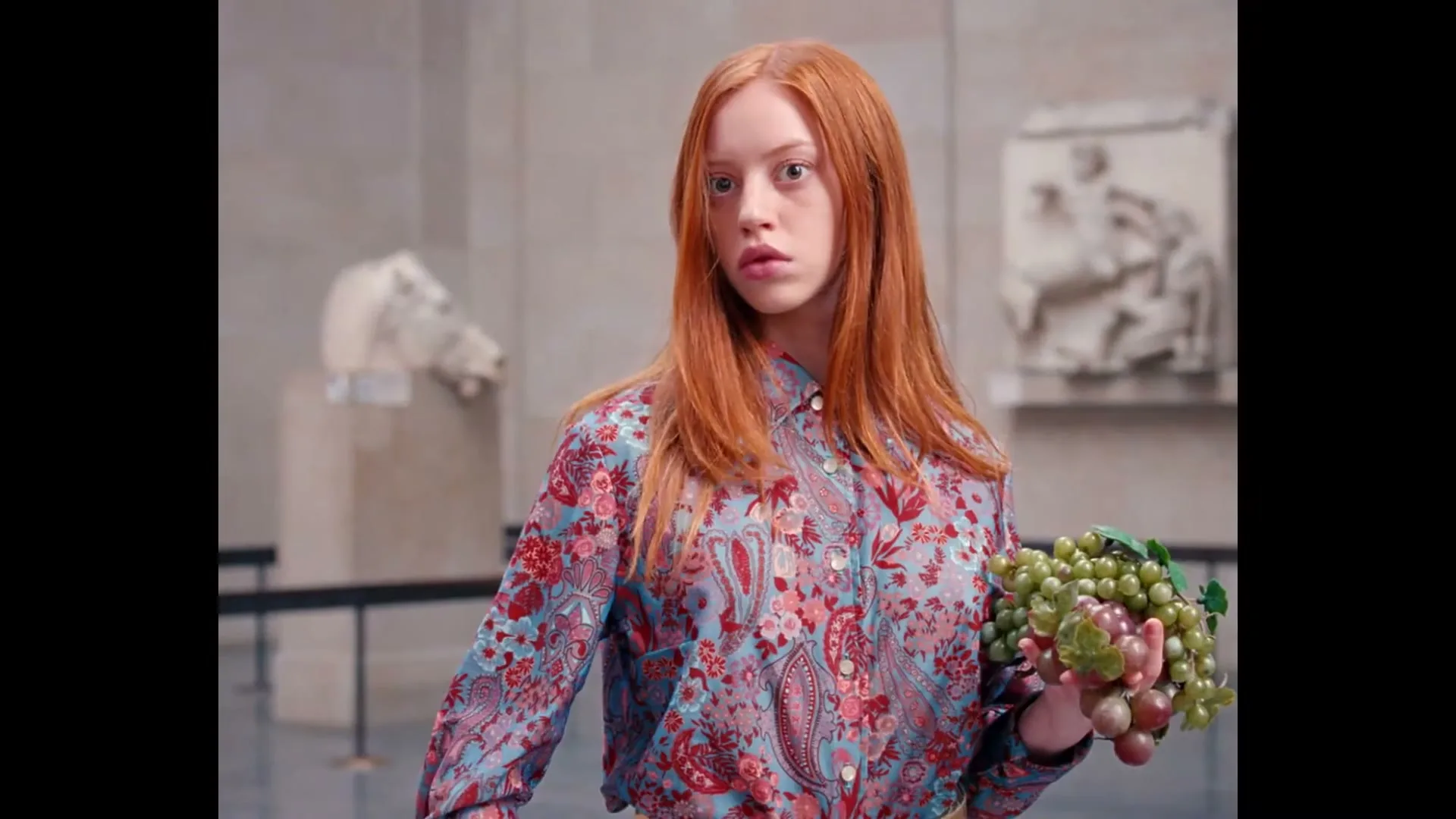A young woman with long red hair, wearing a colorful patterned shirt, holds a bunch of grapes and looks around a museum with ancient sculptures.