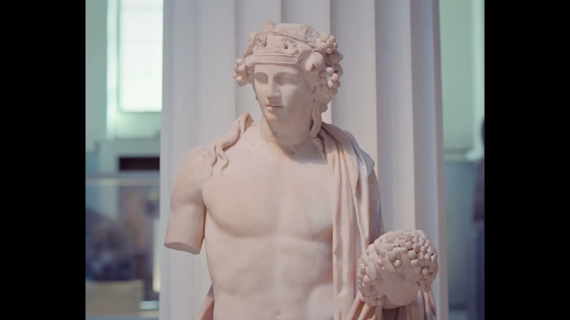 A marble statue of a male figure with a wreath on his head and grapes in his hand is shown in a museum.