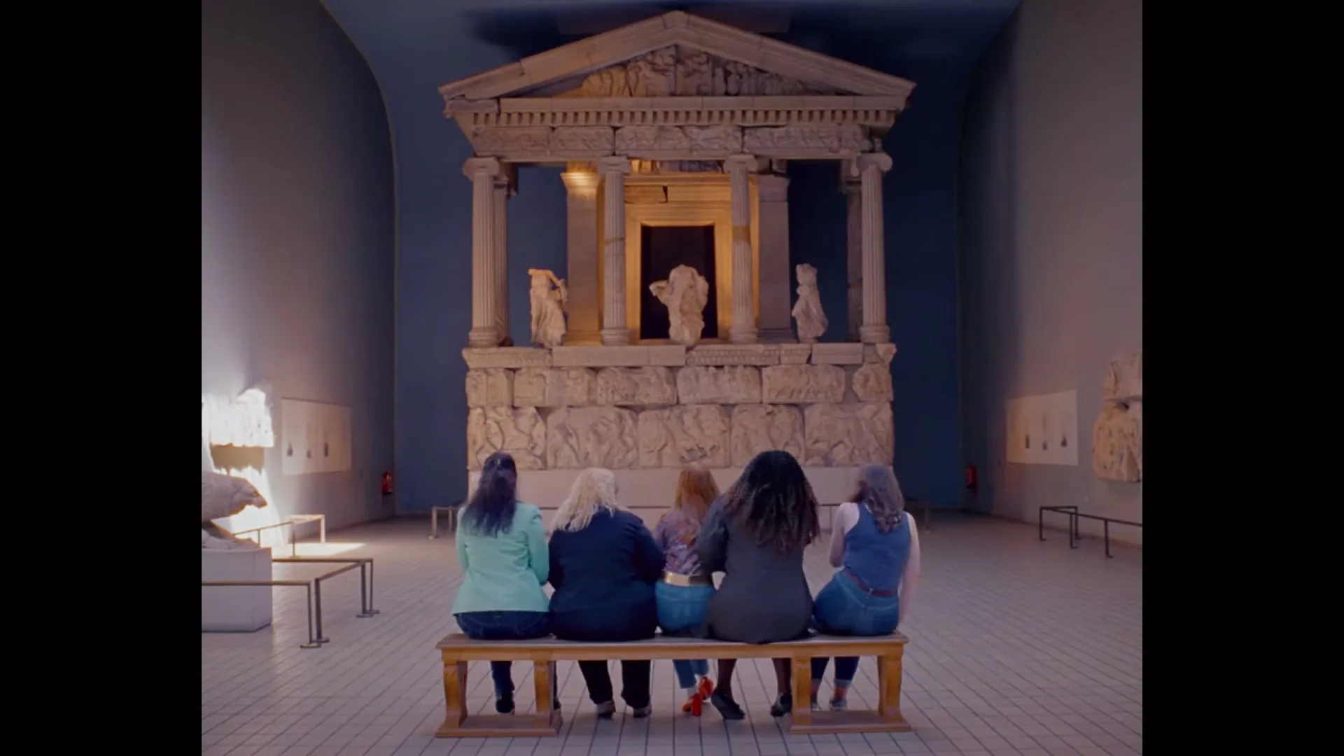 Four women sit on a bench, looking at a large ancient Greek temple exhibit in a museum.