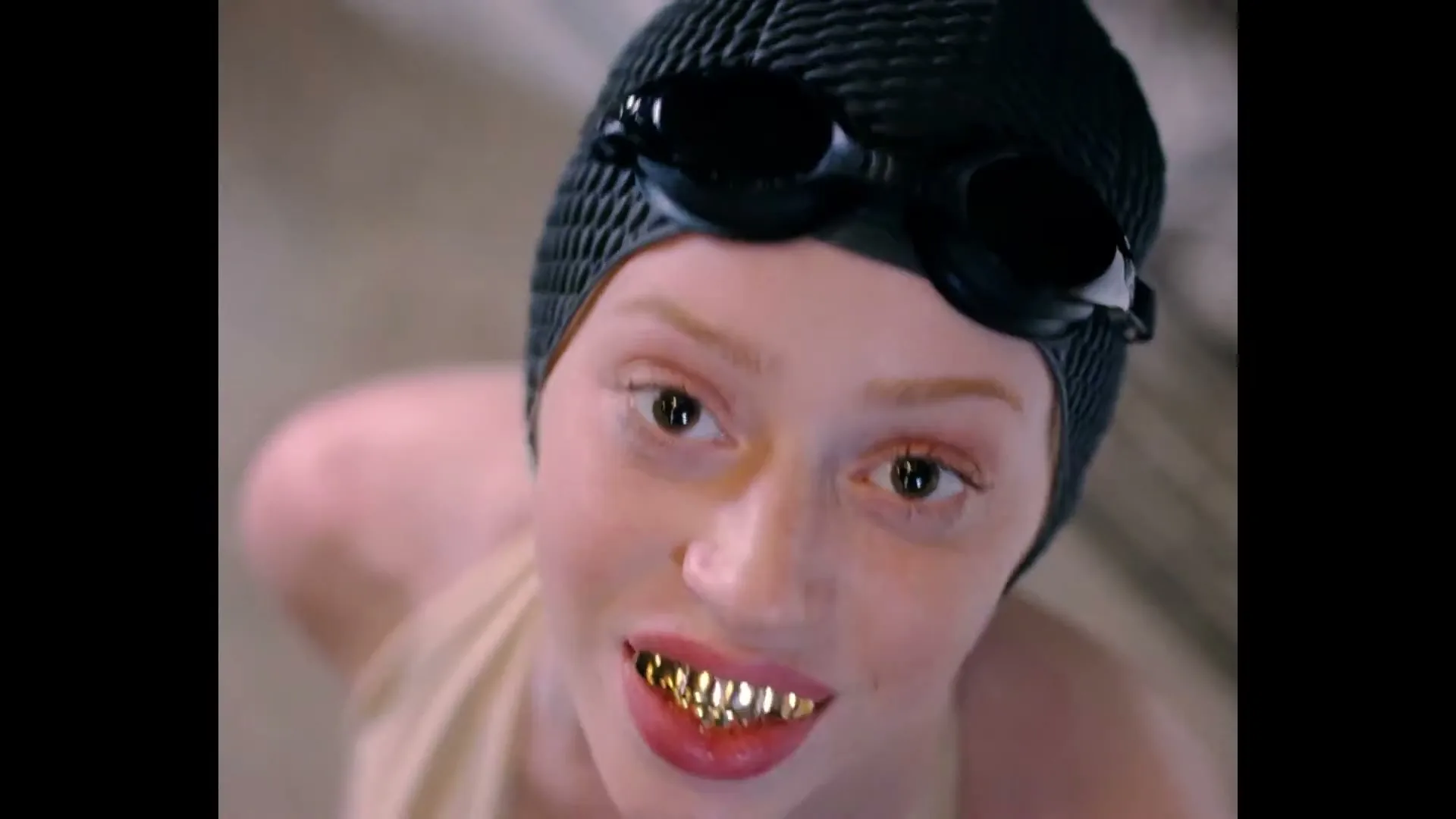A young woman with a black swimming cap and goggles, wearing gold grills, looks up at the camera and then dips her head down, in an indoor swimming pool.