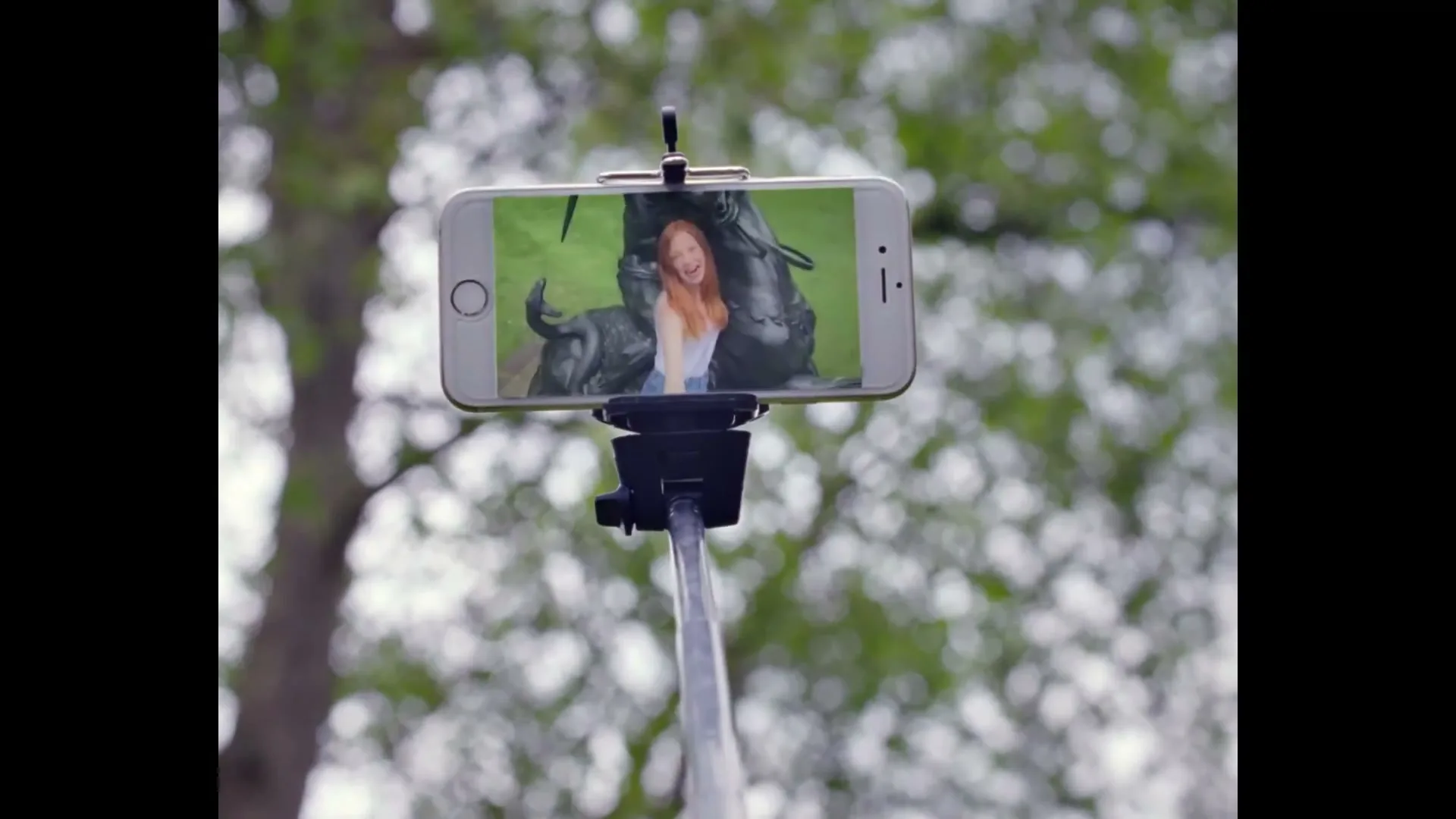 A selfie stick with a smartphone attached is held up against a blurred background of green trees, showing a young woman smiling in a selfie.