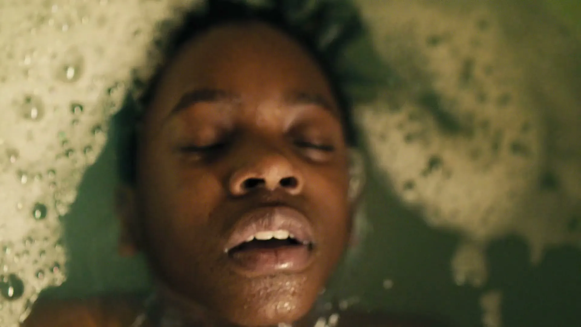 A young Black boy with closed eyes and an open mouth is seen from above, floating in a bubble bath.