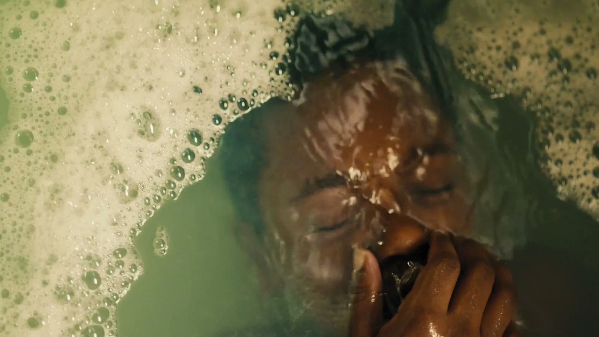 A person with dark skin and closed eyes is submerged in a bubbly, green-tinted bath, with water rippling over their face.