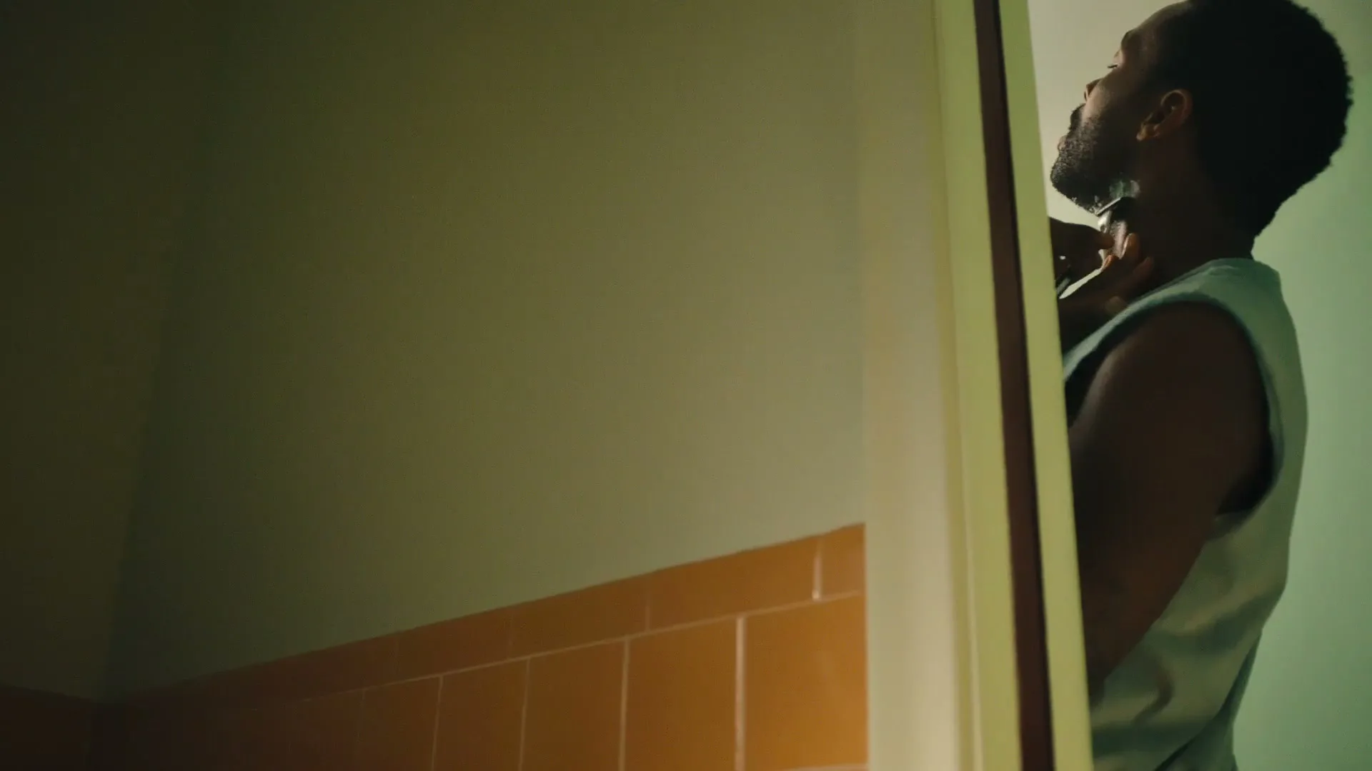 A man with a beard shaves his neck in a bathroom, seen through a slightly ajar door.