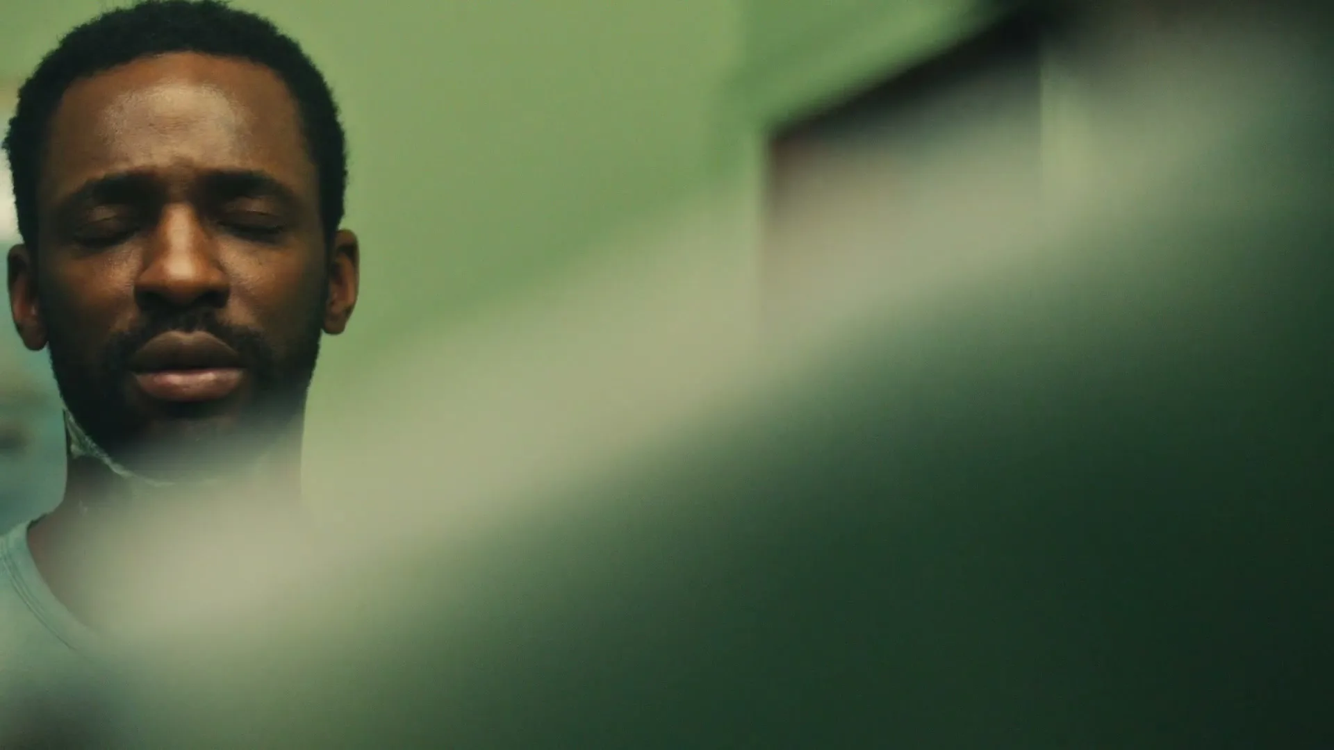 A Black man with a beard and short hair opens his eyes and looks up, partially obscured by a blurry foreground, in an interior setting.