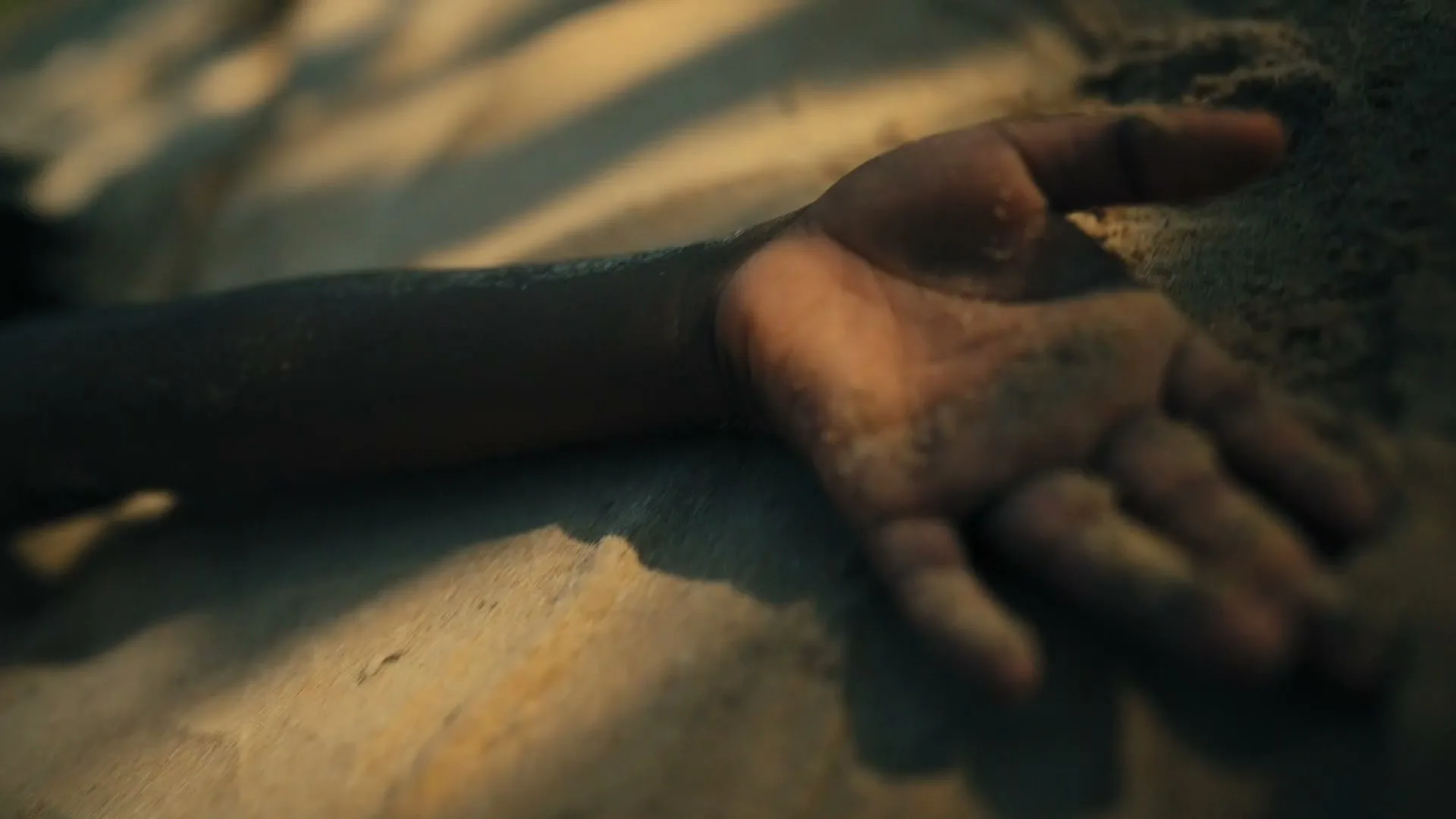 A child's arm and hand, covered in sand, rests on a sandy surface in soft, natural light.