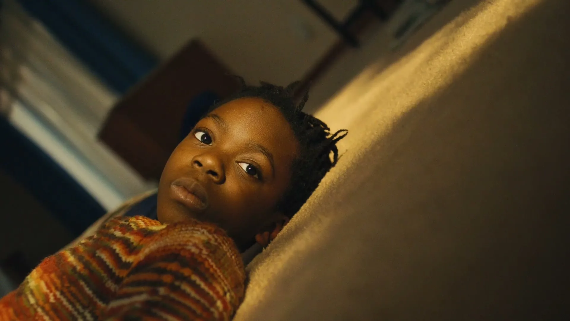 A young Black child with dreadlocks lies on a couch, looking directly at the camera with a neutral expression in a dimly lit room.