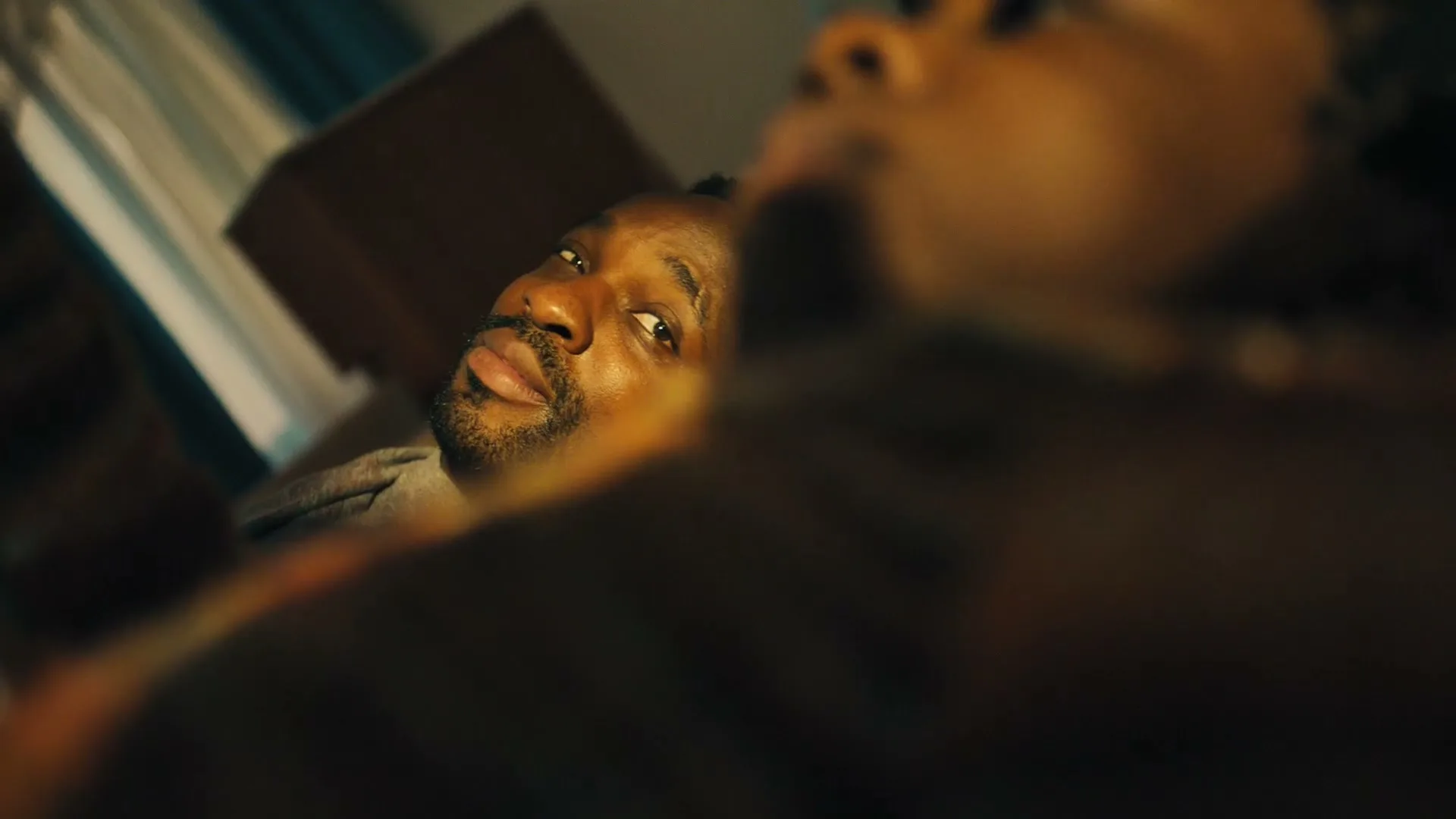 A man with a beard lies in bed, looking up and to the right, with another person out of focus in the foreground.