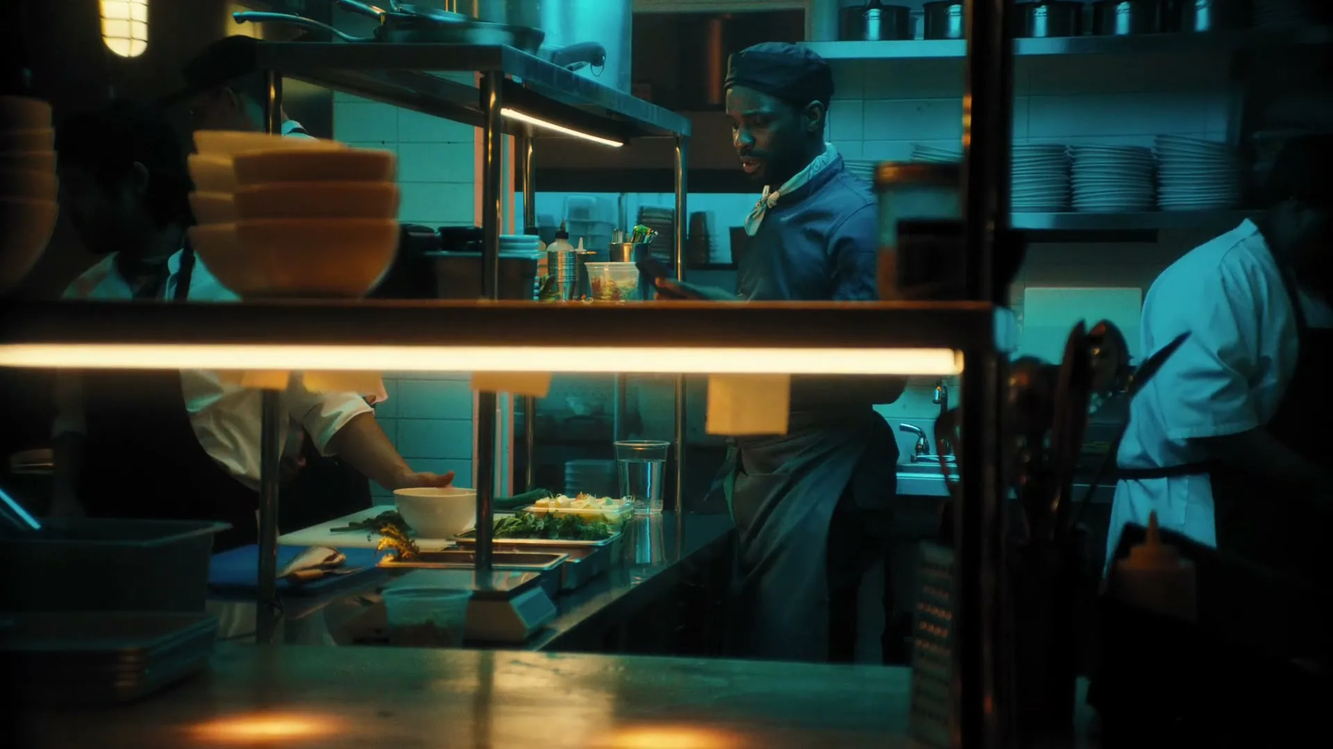 A male chef in a blue uniform and black cap works in a busy, dimly lit kitchen, preparing food at a counter.