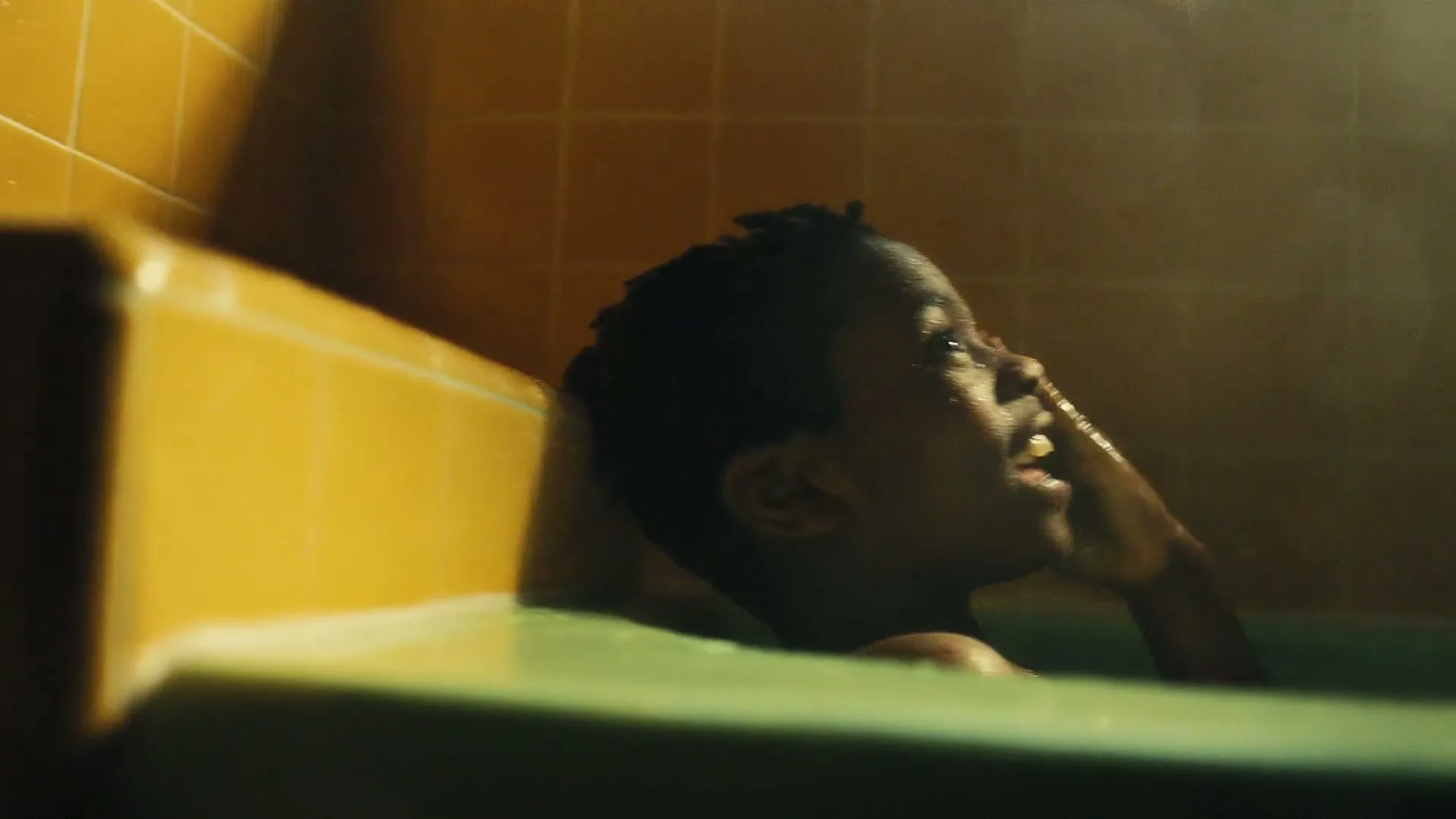 A young Black boy with short dark hair is seen from the side, sitting in a bathtub with green water, looking up and opening his mouth as if catching water.