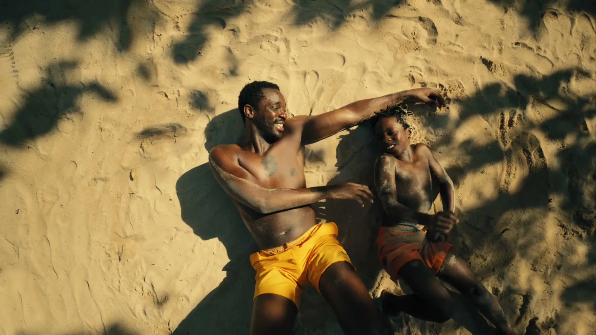A man and a child, both shirtless and wearing swim trunks, lie on a sandy beach, laughing and playing in the sand under dappled sunlight.