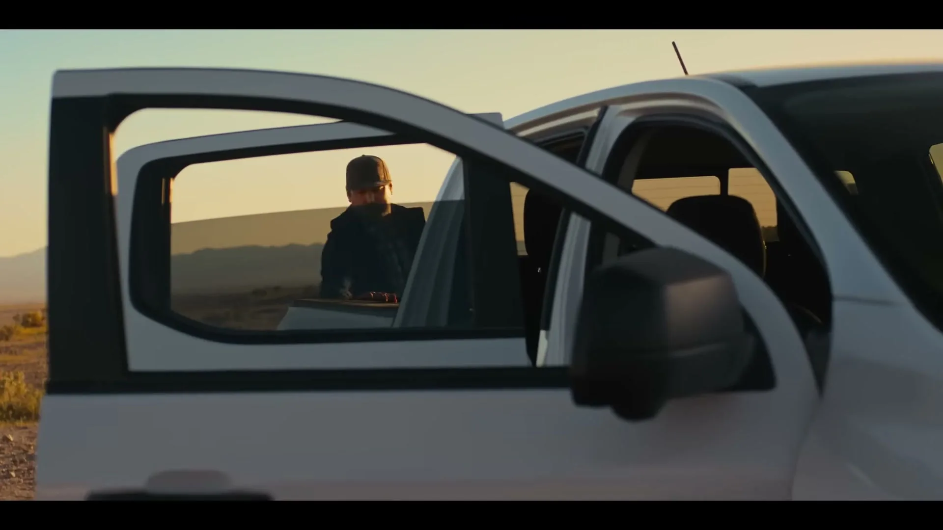 A man with a beard and cap stands by a white Ford Ranger truck with its door open, looking out at a vast, arid landscape at sunset.