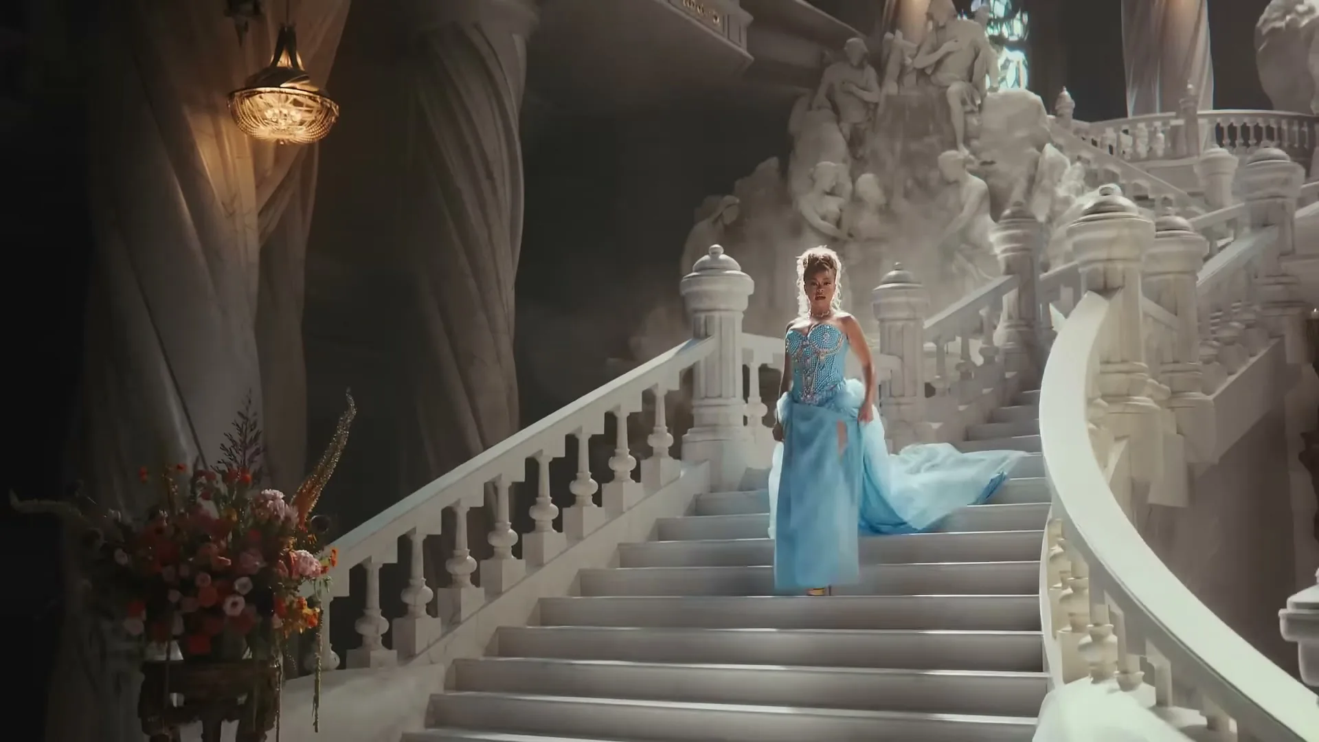 A woman in a blue gown descends a grand white staircase in a lavish, ornate hall, with a large floral arrangement in the foreground.