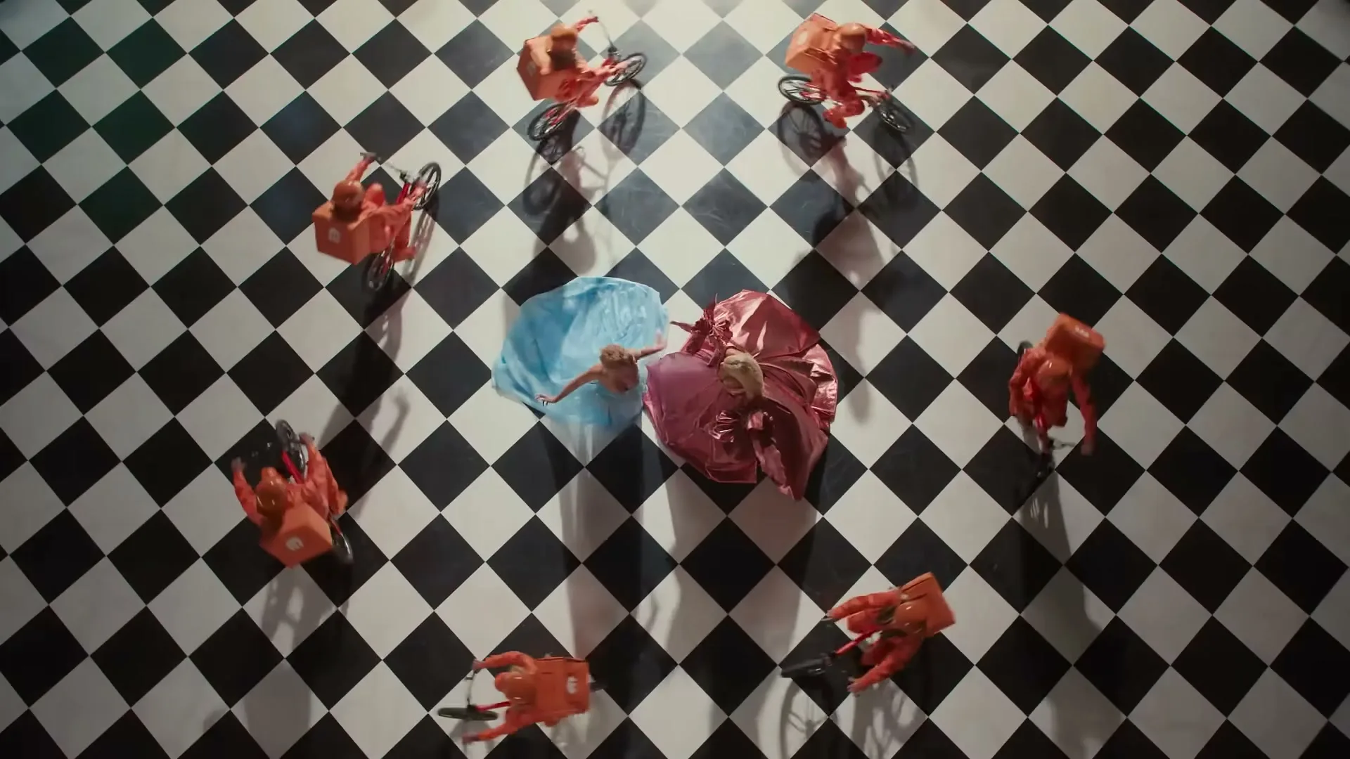 Two women in flowing dresses dance on a black and white checkered floor, surrounded by several delivery riders on bicycles.