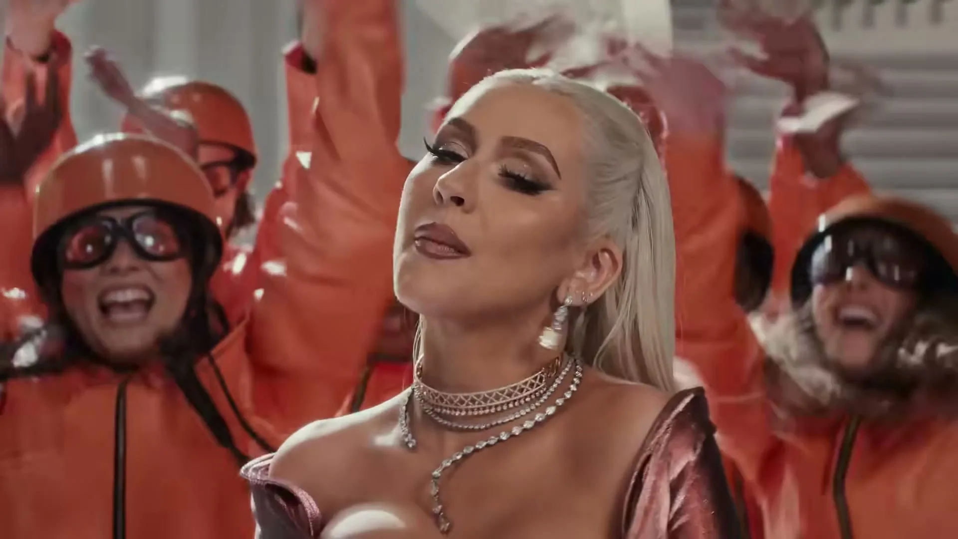 Christina Aguilera, a blonde woman in a sparkly dress, sings with a group of people in orange jumpsuits behind her in a grand hall.