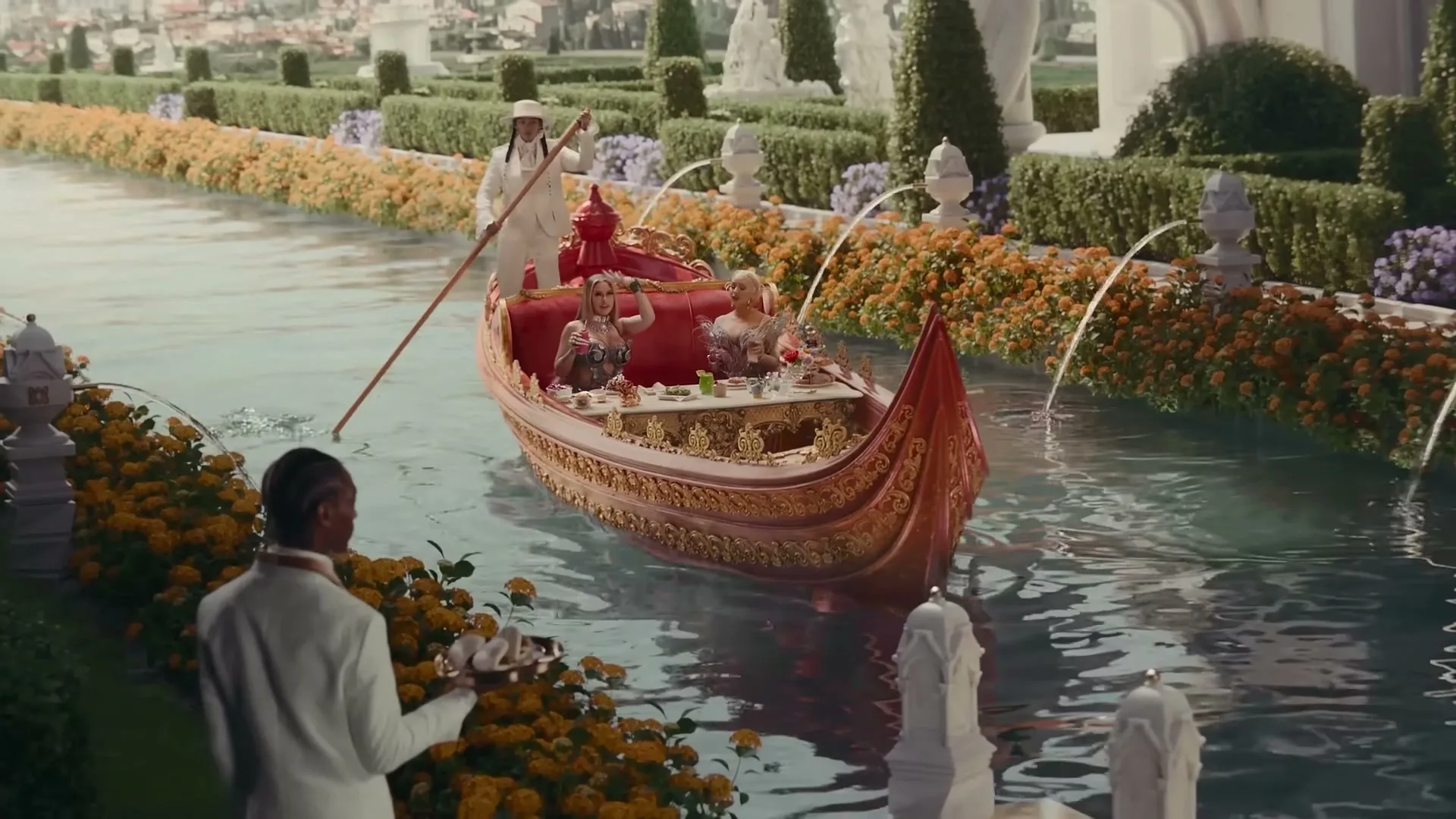 A gondola with two women in elaborate outfits is punted down a canal by a gondolier, while a waiter stands on the bank.