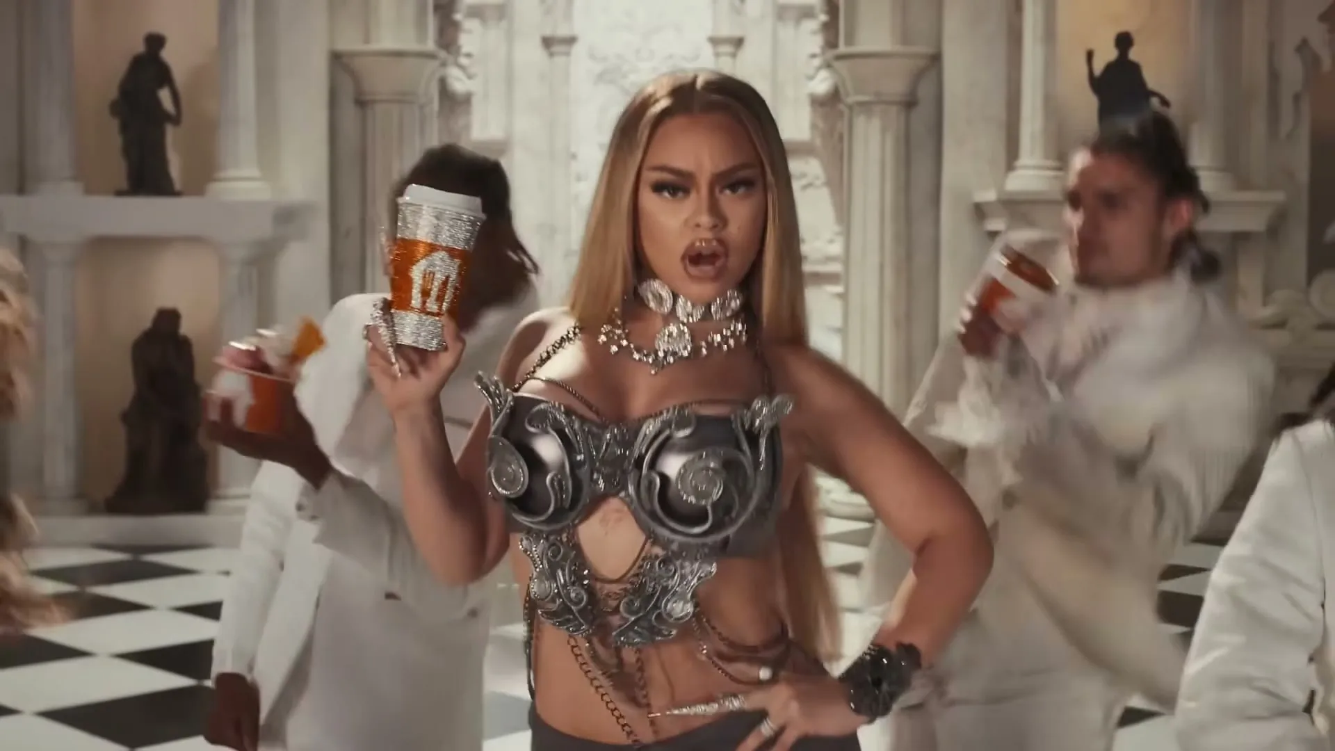 Latto, a light-skinned woman with long blonde hair, wearing a silver bra and skirt, holds a coffee cup and sings while dancing with a group of people in a grand hall.