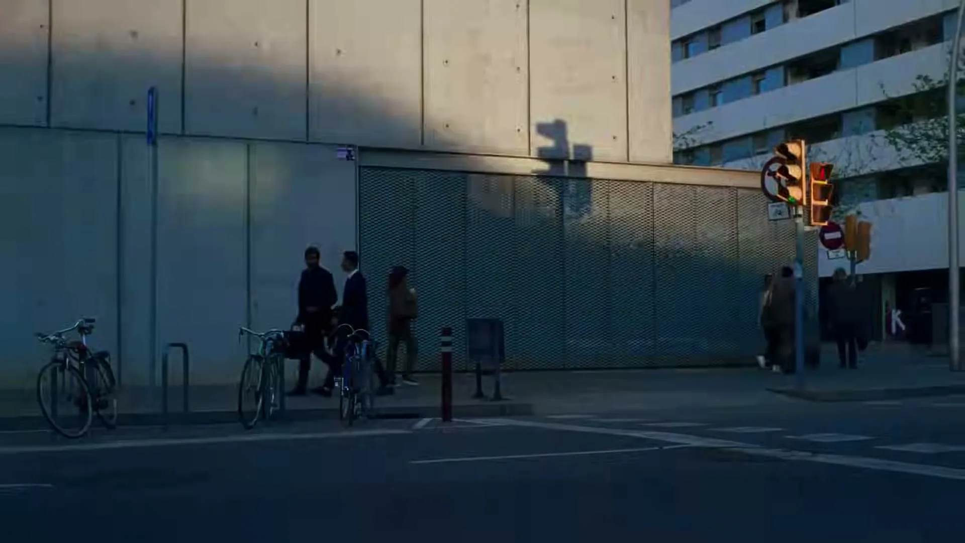 A wide shot of a city street corner with pedestrians walking, bicycles parked, and a traffic light changing, under the warm glow of the sun.