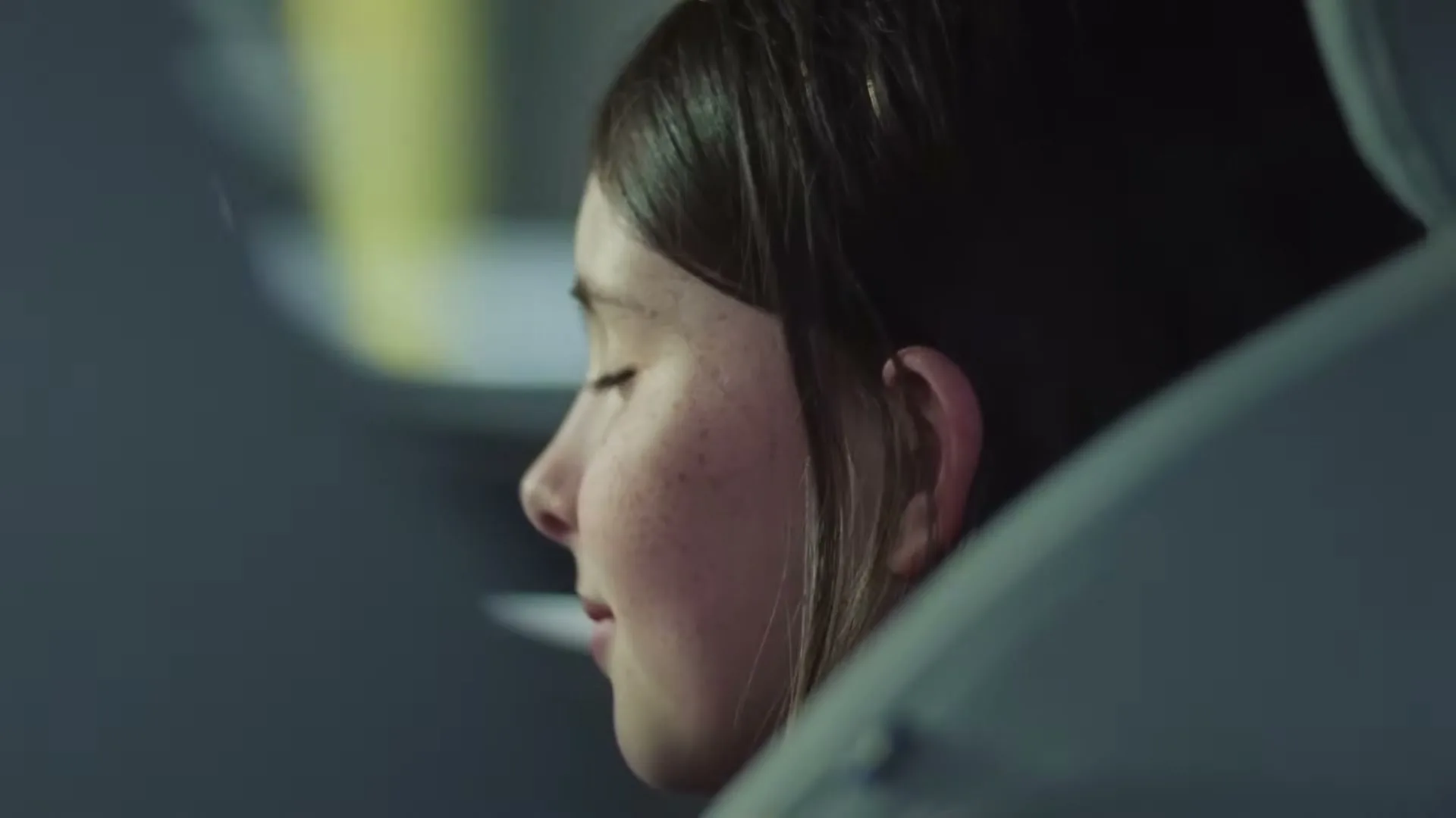 A young girl with dark hair and freckles sits in a car, looking out the window with a slight smile.