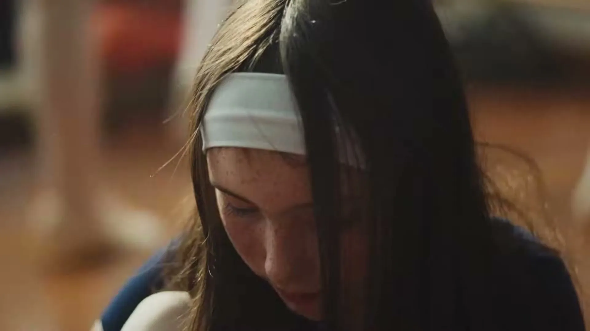A young girl with long dark hair and a white headband looks down, then slowly lifts her gaze, in a warm, softly lit interior.