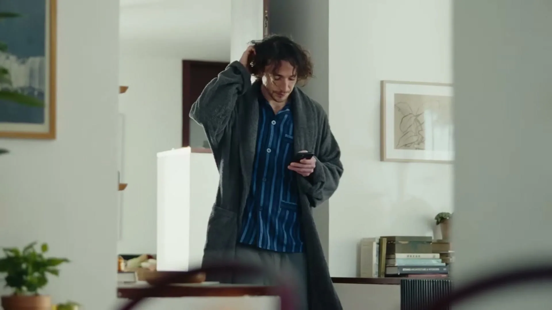 A young man with messy hair, wearing pajamas and a robe, stands in a bright living room, looking at his phone and scratching his head.