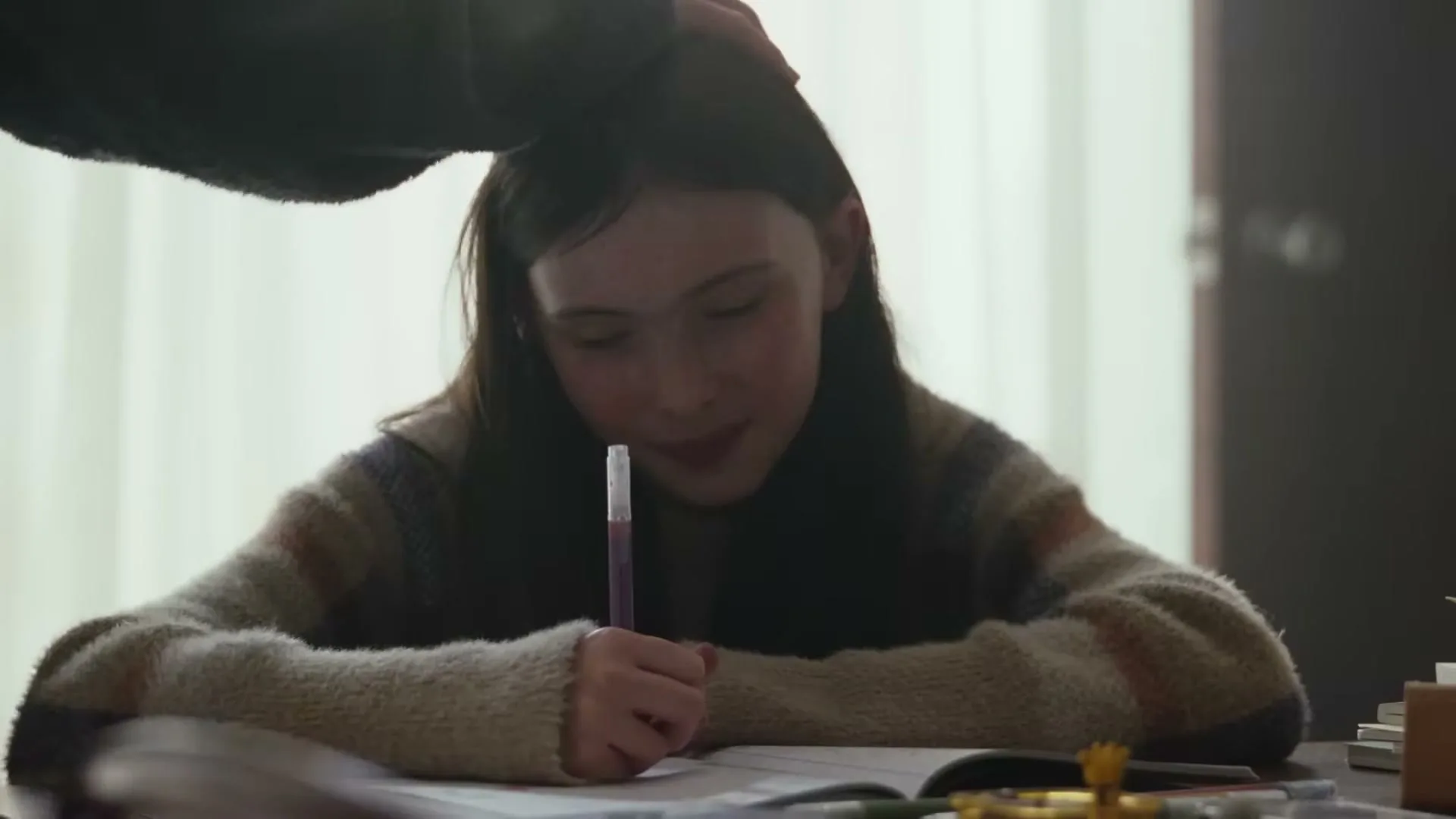 A young girl with long dark hair, wearing a striped sweater, sits at a desk and writes in a notebook while an adult hand gently pats her head.