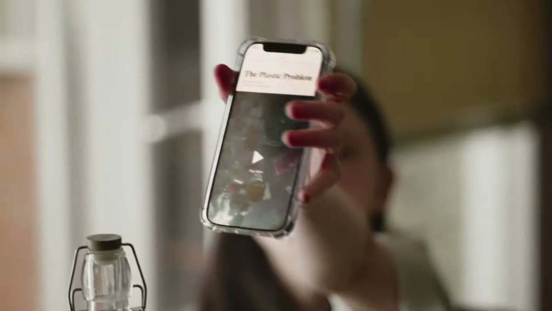 A young woman with long dark hair holds up a smartphone to show the screen, which displays a video titled "The Plastic Problem."