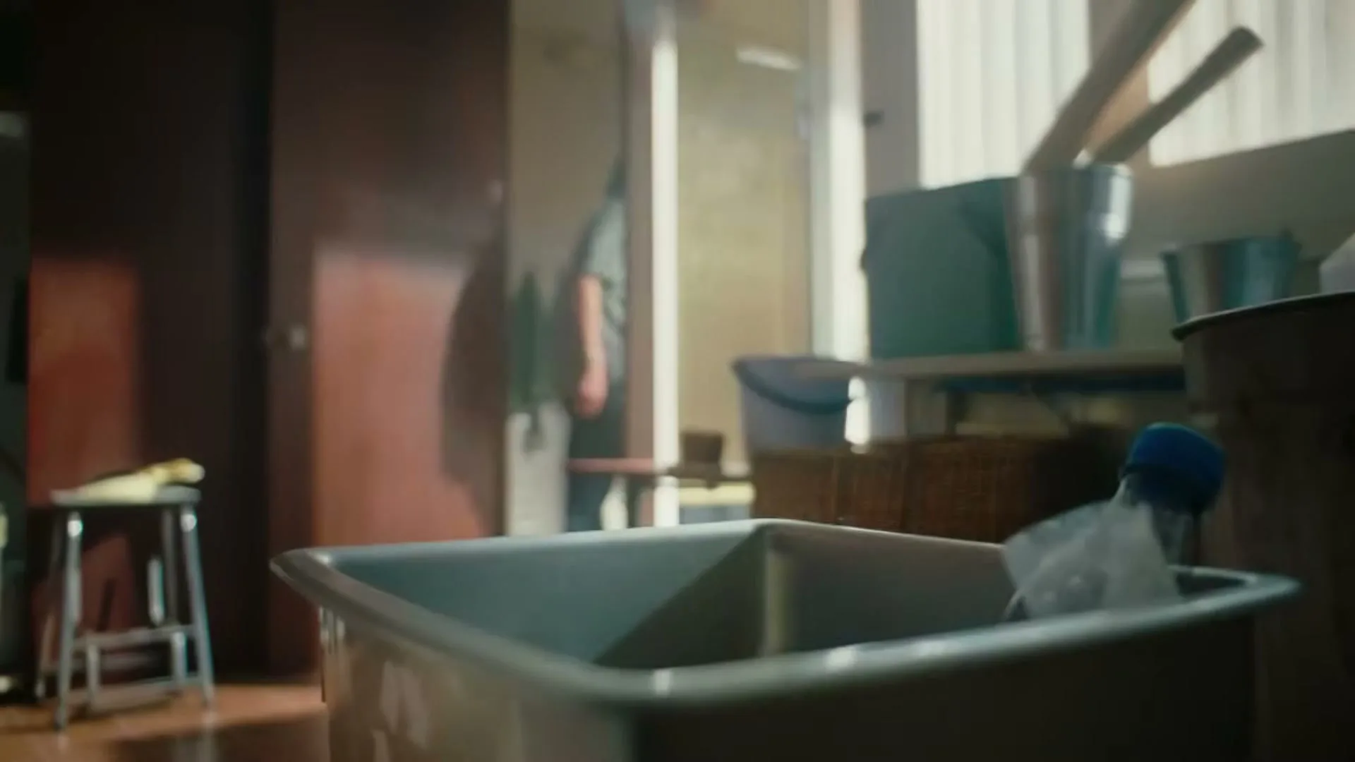 A young man walks into a utility room and throws a plastic bottle into a recycling bin, then walks out.