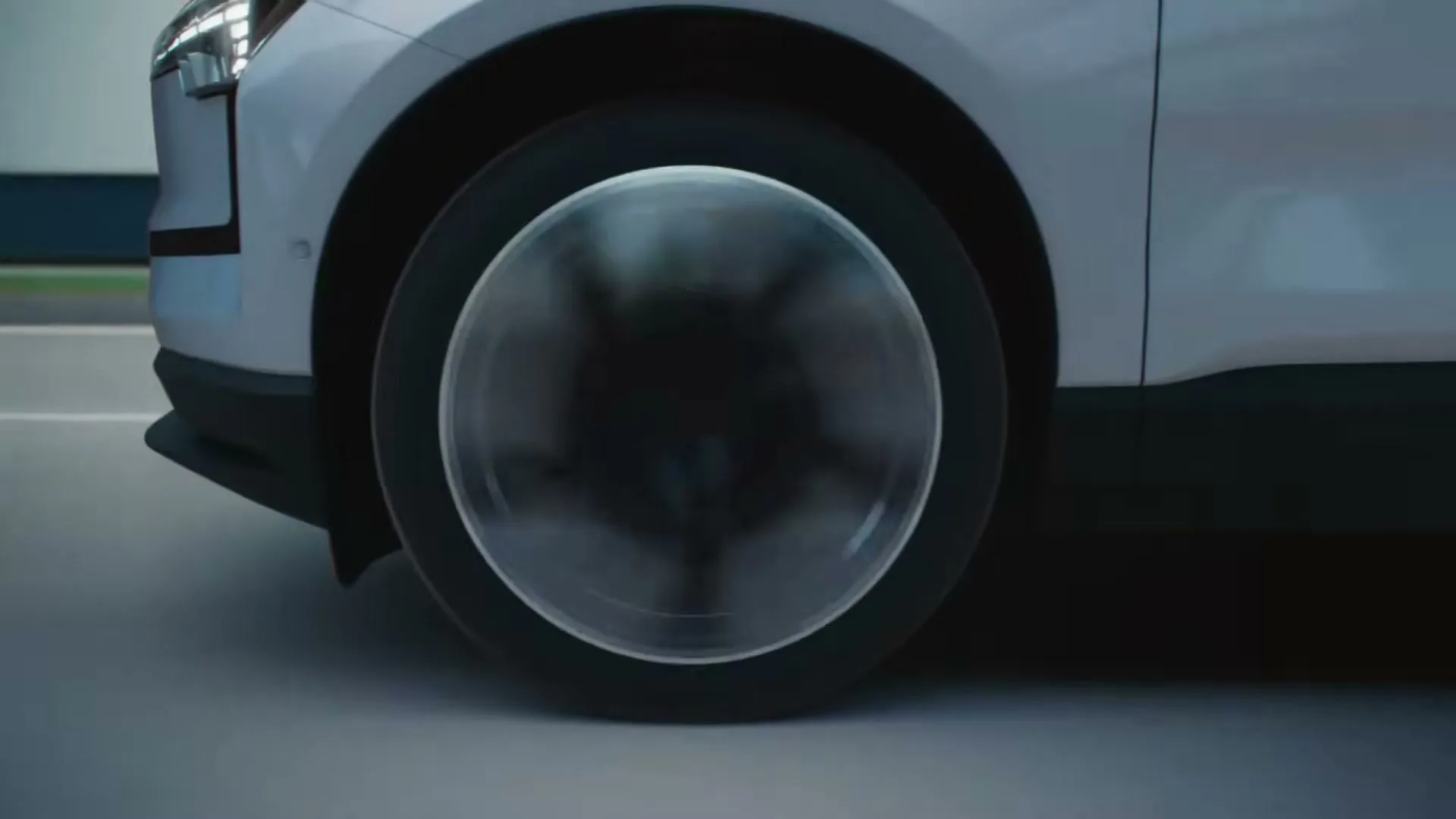 A white car's front wheel with a clear hubcap spins as the vehicle drives on a road.