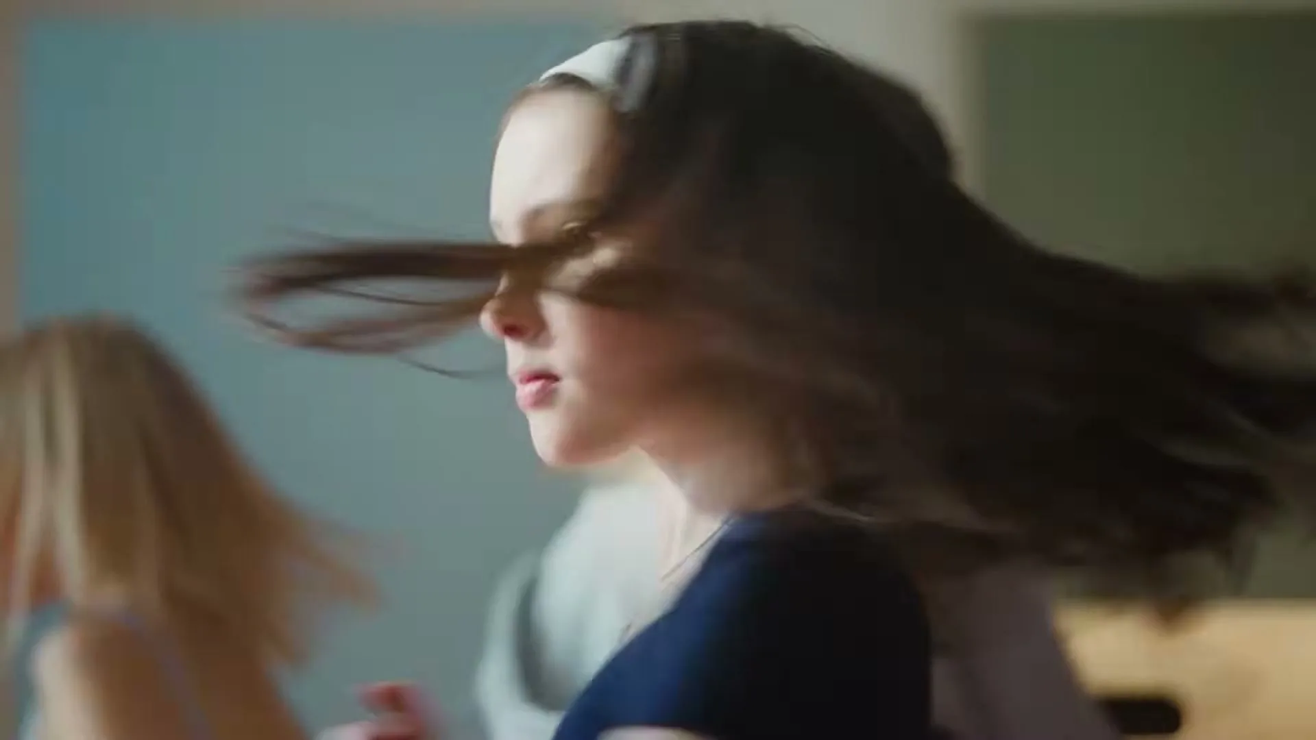 A young girl with long brown hair and a white headband turns around, her hair flowing, as two other girls are blurred in the background.
