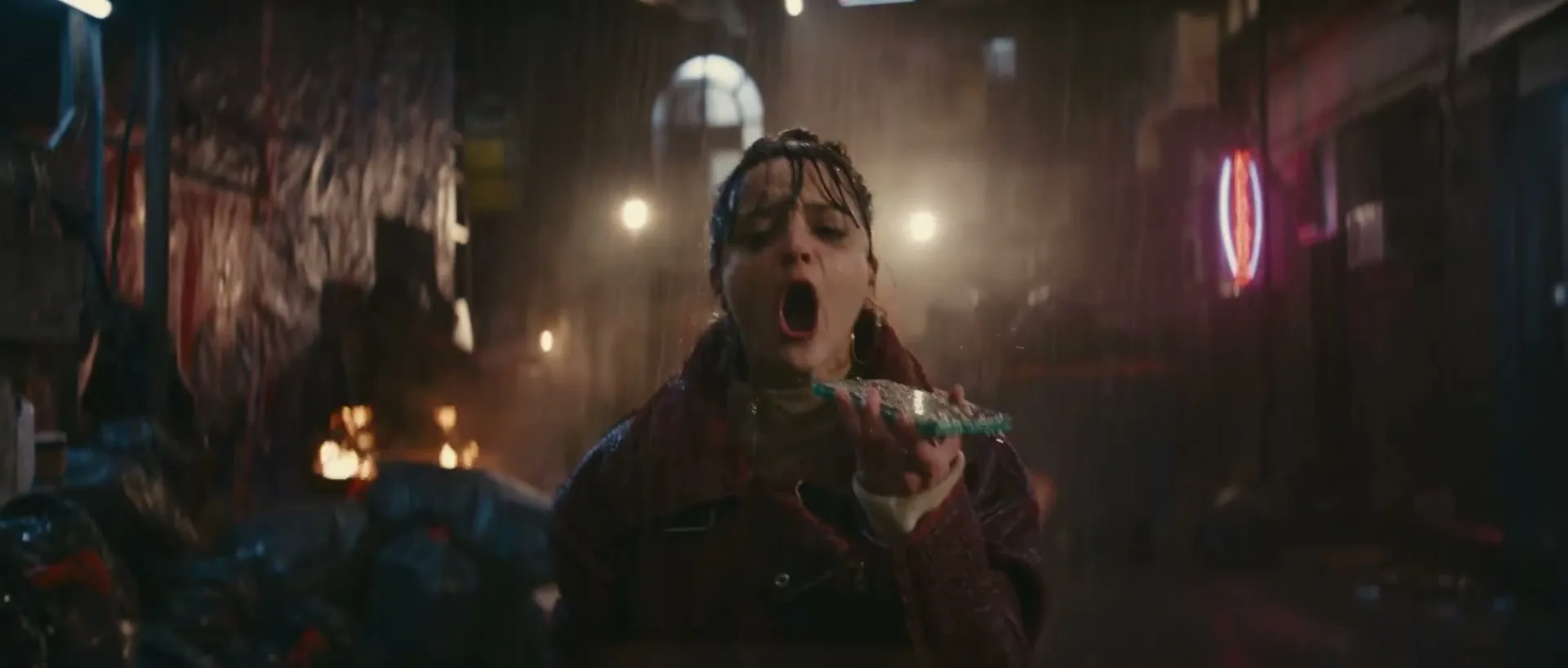 A young woman with dark hair in a bun, wearing a brown jacket, shouts into a green object in a dark, rainy alleyway with fires burning.