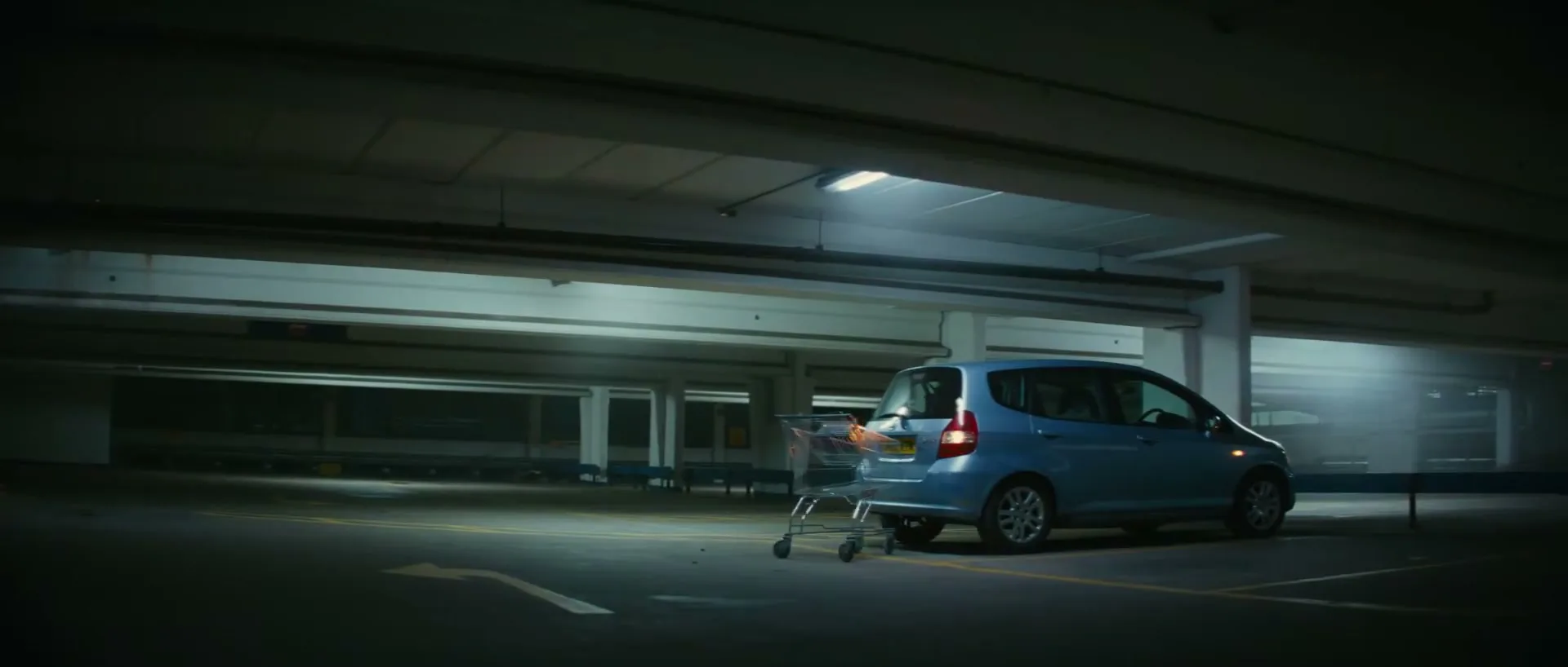 A light blue car is parked in a dark, empty underground parking garage, with a shopping cart rolling into its rear bumper.
