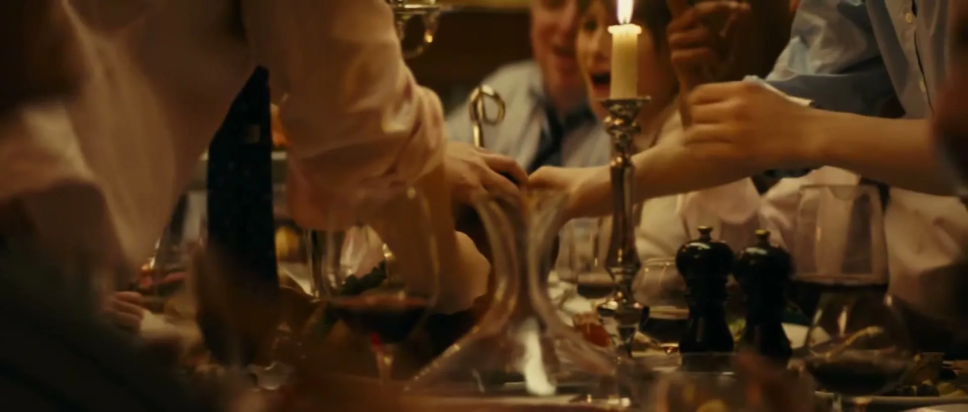 A group of people at a dinner party are seen from a low angle, reaching for a decanter of red wine on a candlelit table.