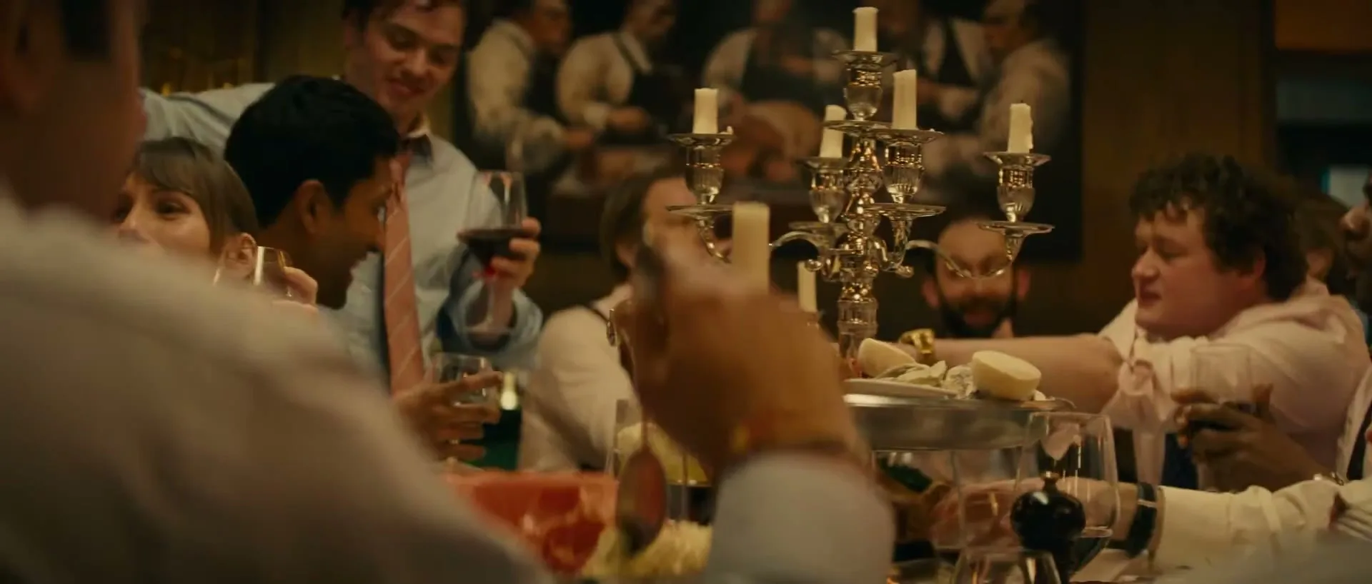 A diverse group of people in formal wear are seated around a large dining table, eating and drinking, with a candelabra in the center.