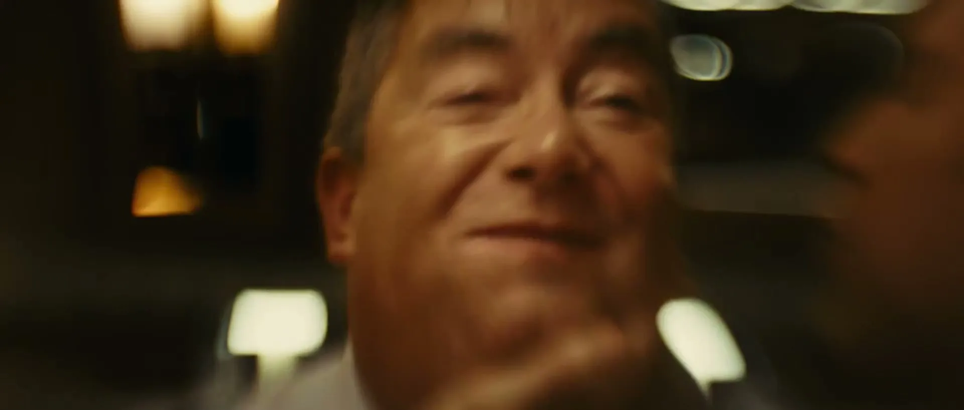 A middle-aged man with grey hair and a white shirt smiles broadly, revealing crooked teeth, in a dimly lit interior with bokeh lights.