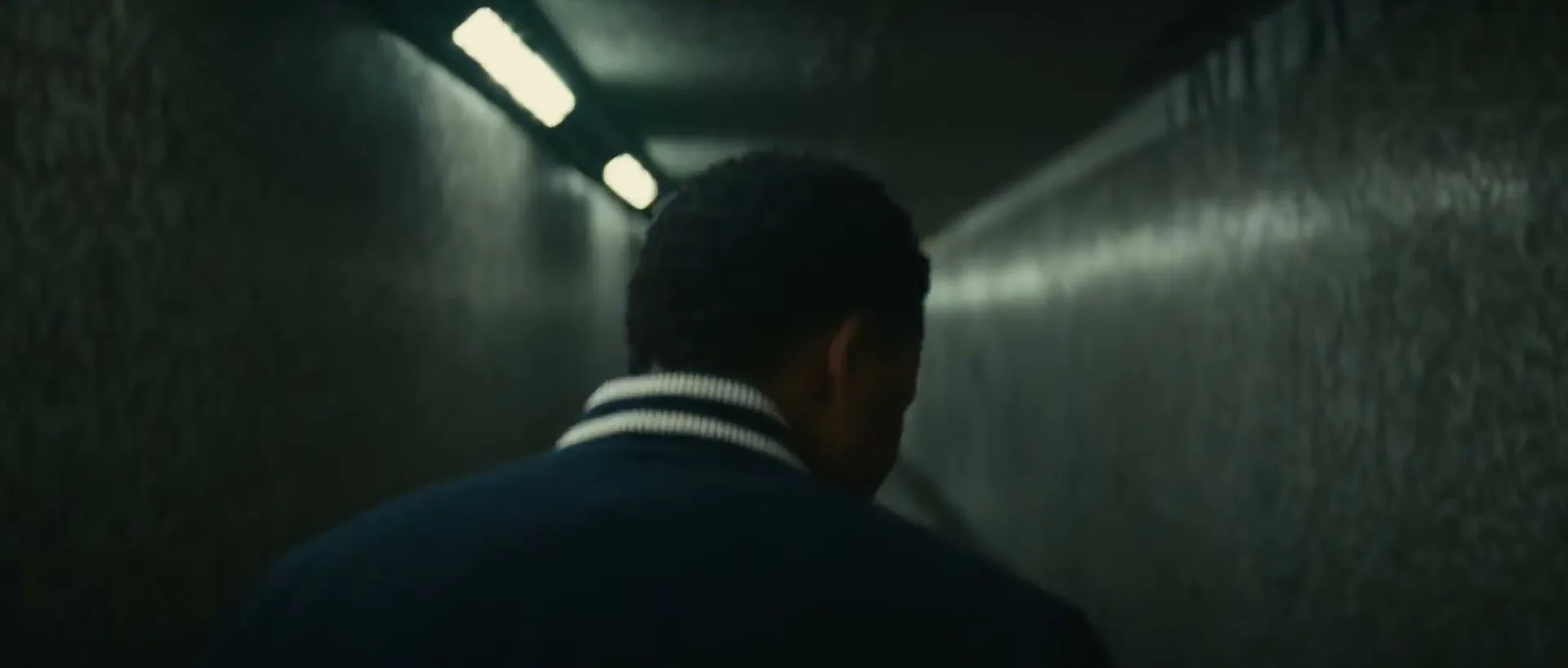A young man with dark curly hair, wearing a dark blue jacket with white striped collar, walks away from the camera down a dimly lit, narrow concrete tunnel.