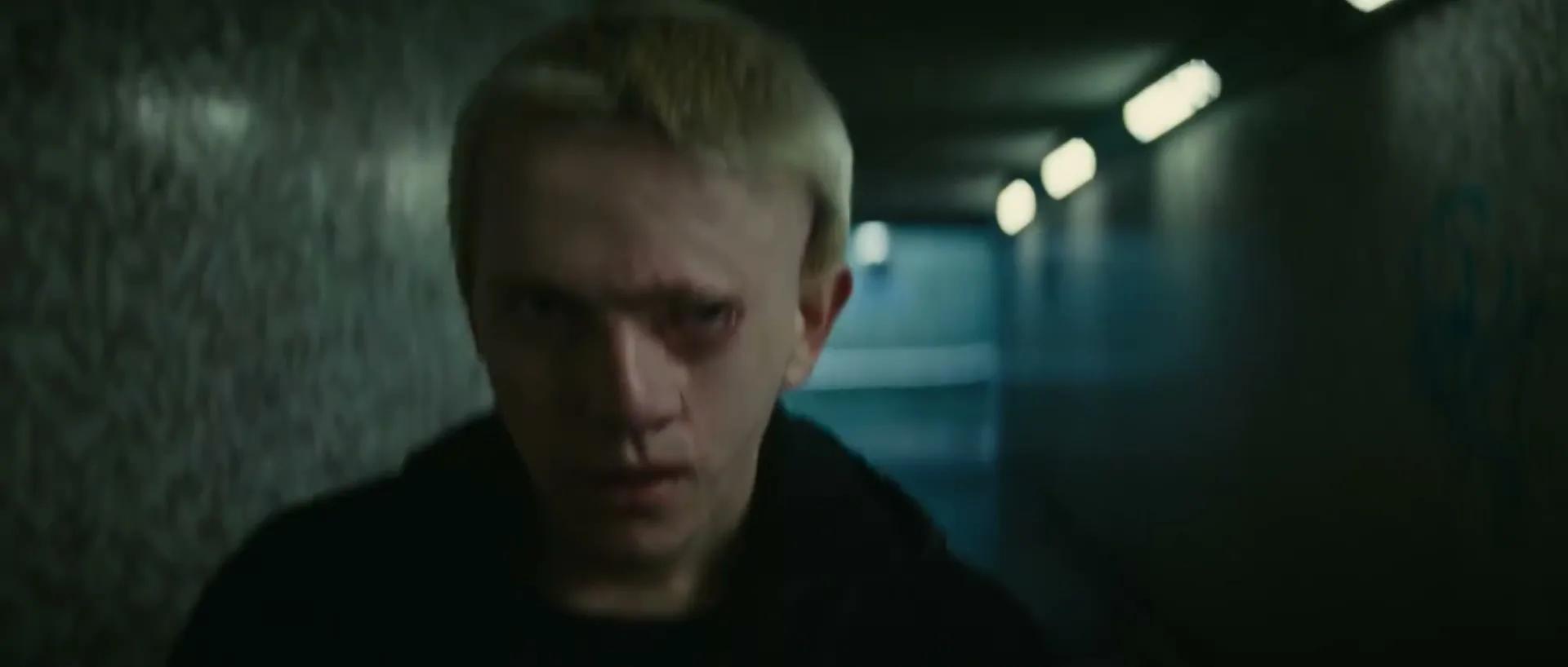 A young man with blonde hair and a black hoodie looks intensely at the camera in a dimly lit, concrete tunnel.
