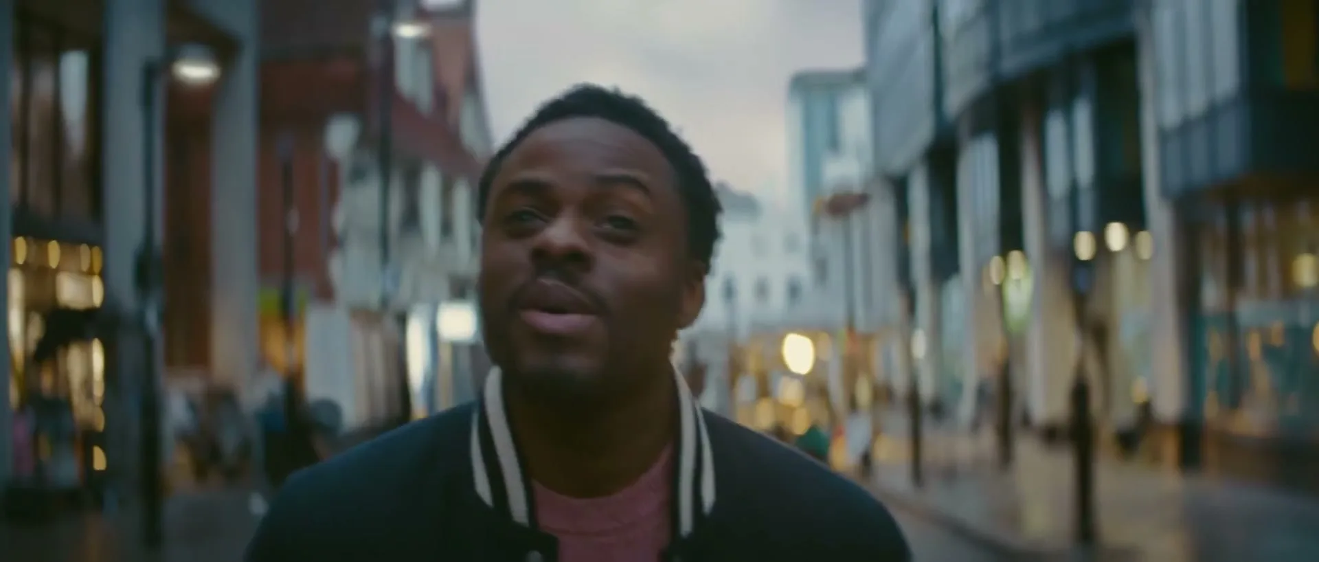 A young Black man in a varsity jacket walks down a city street at dusk, looking around with a thoughtful expression.
