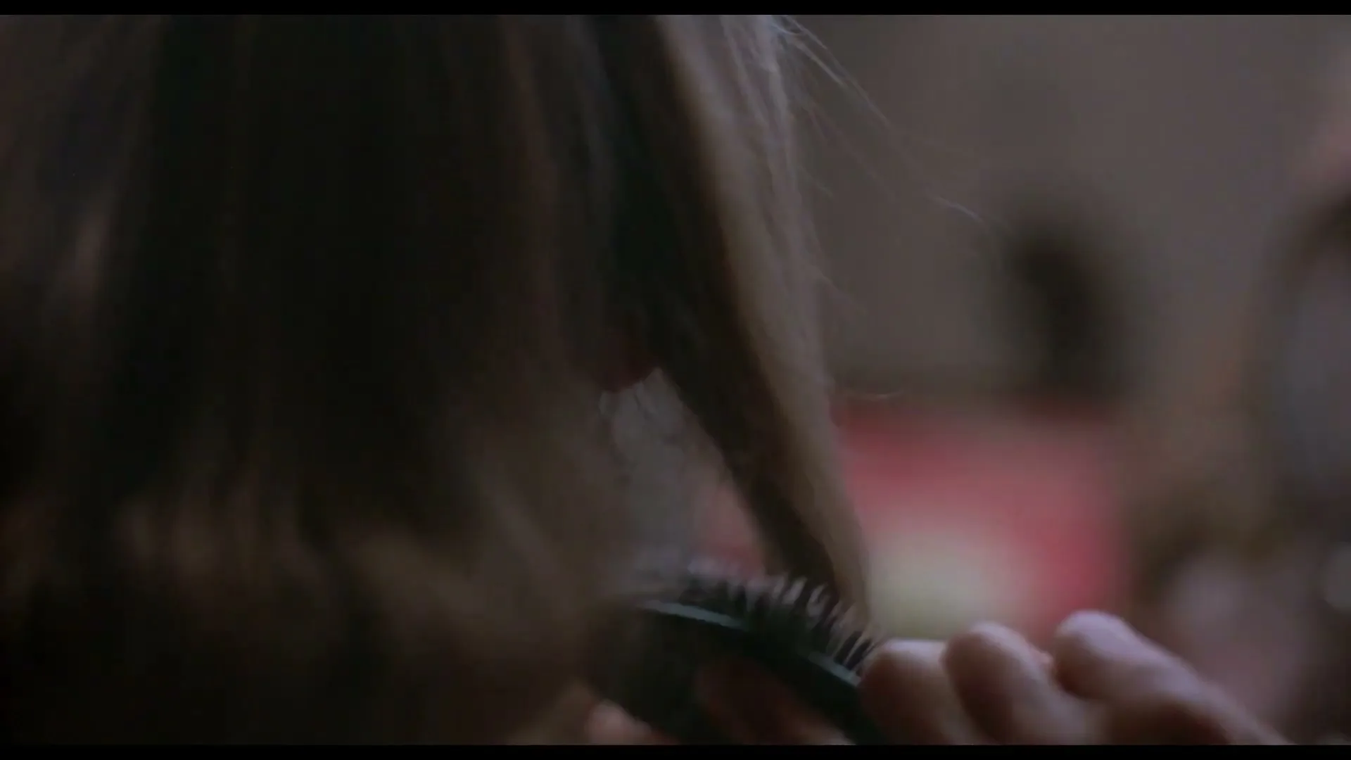 A close-up shot shows an older woman with long blonde hair being gently brushed by another person's hand, in a dimly lit interior.