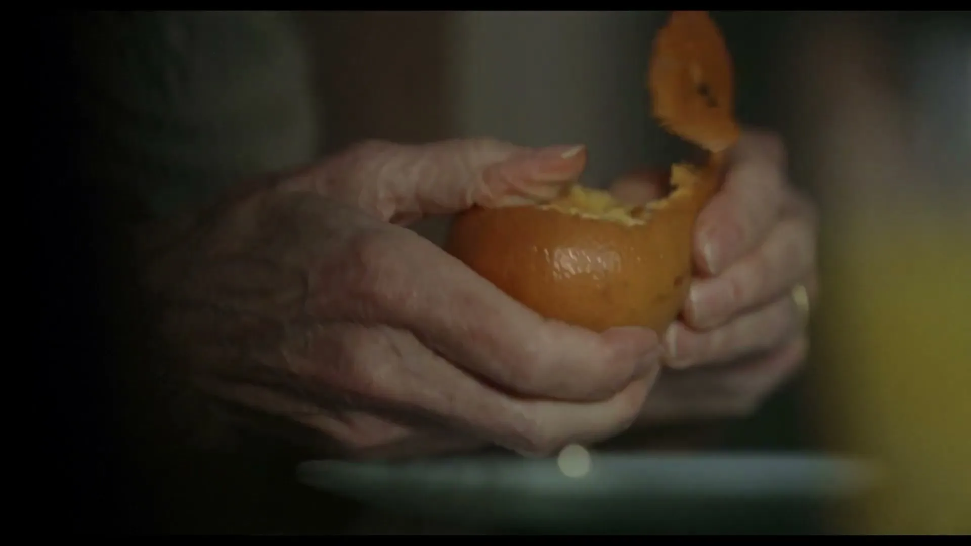 Wrinkled hands of an older person struggle to peel an orange, revealing the fruit underneath, in a dimly lit interior.