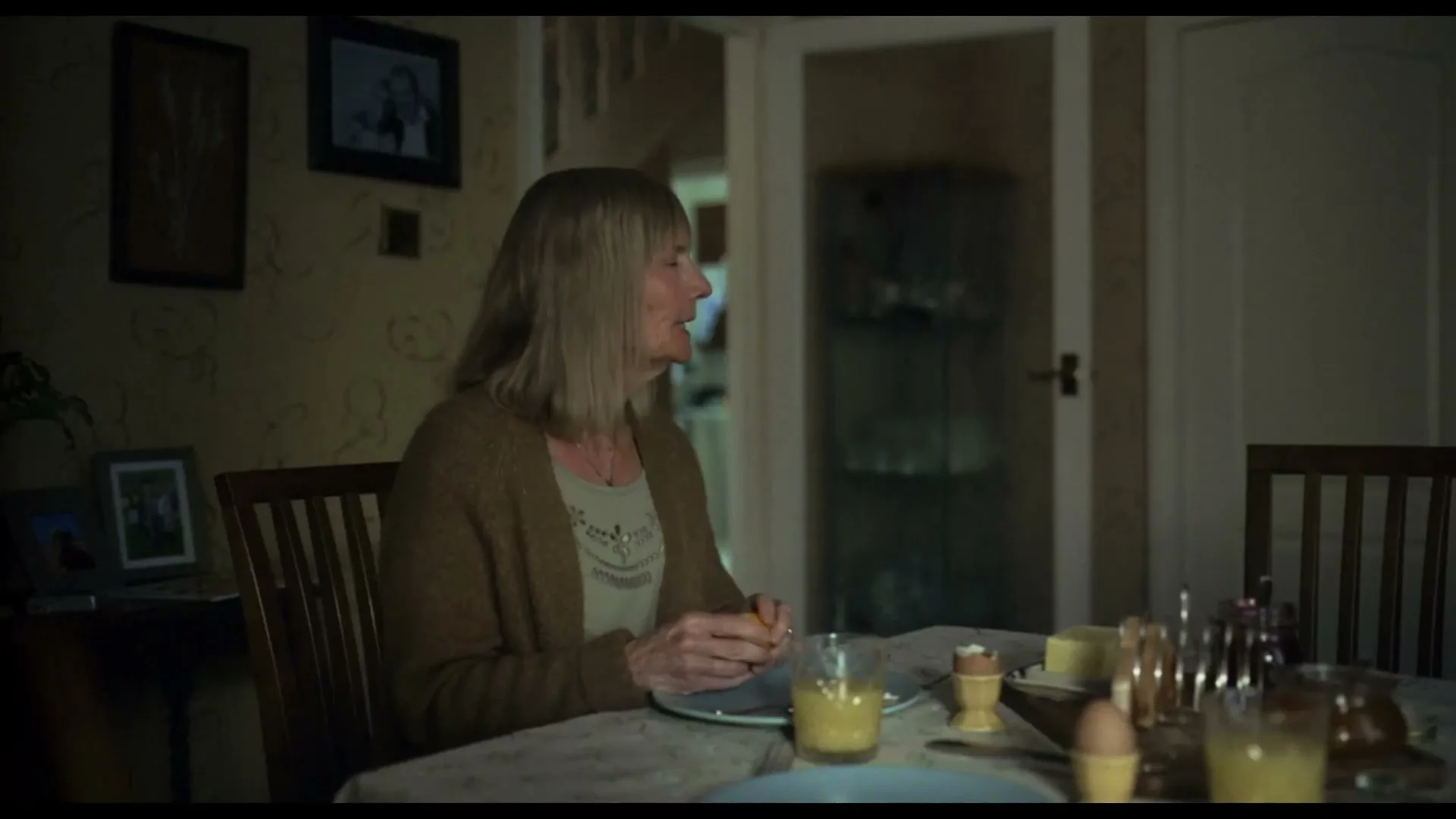 A senior woman with blonde hair, wearing a brown cardigan, sits at a dining table, peeling an orange, in a dimly lit dining room.