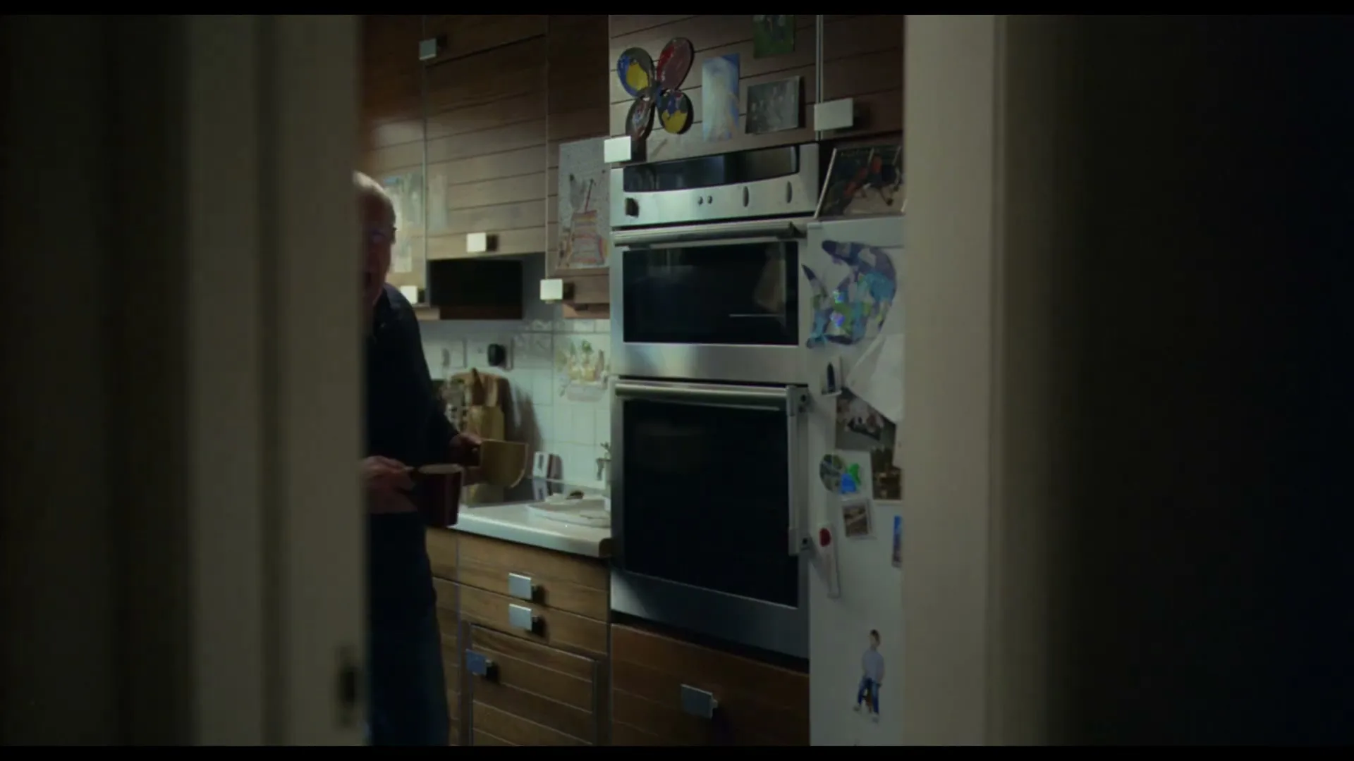 An older man with white hair and glasses, wearing a dark blue sweater, stands in a kitchen holding two mugs, looking confused.