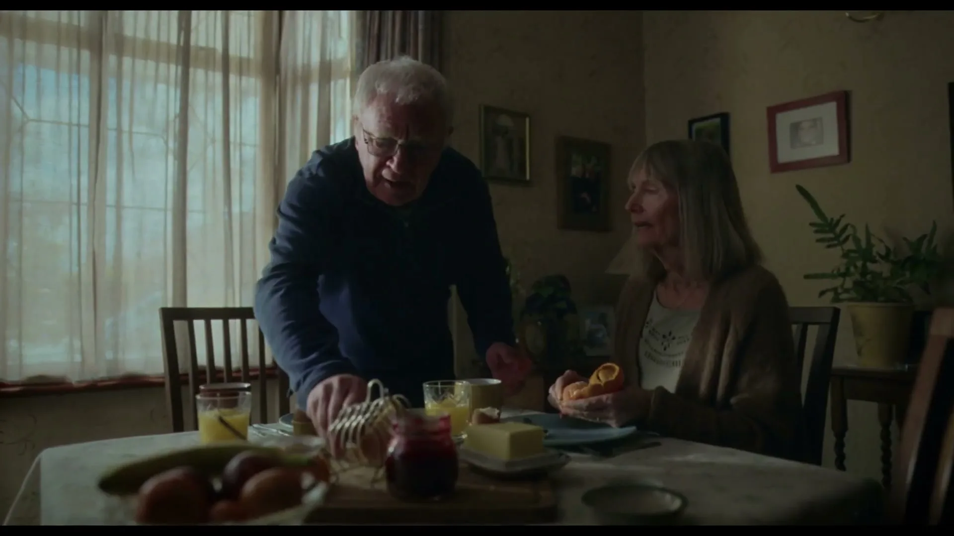An elderly man in a blue sweater and glasses stands over a breakfast table, talking to an elderly woman in a brown cardigan who is peeling an orange.