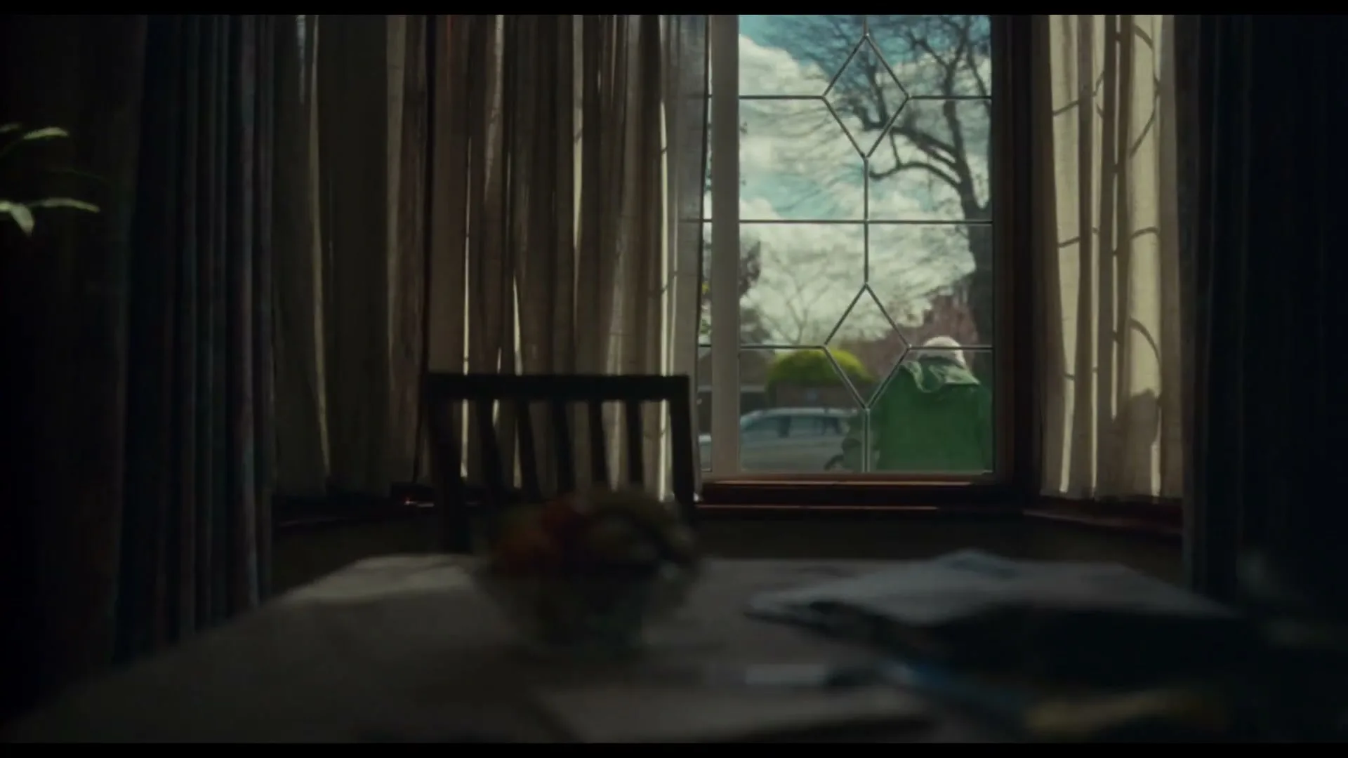 A wide shot from inside a dark room shows a table and a window, through which two people in raincoats walk past a house on a sunny day.