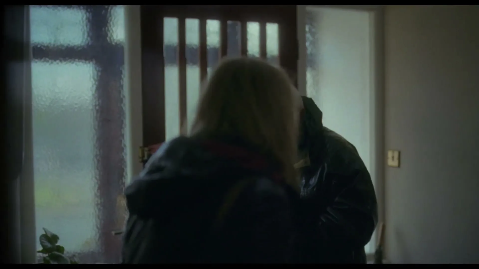 A woman with blonde hair and a man in a dark jacket stand in a dimly lit hallway, talking by a front door with frosted glass panels.