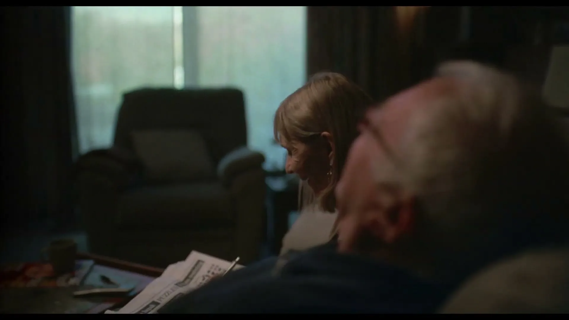An older man with white hair sits on a couch, seen from behind, while a woman with blonde hair sits beside him, smiling, in a dimly lit living room.