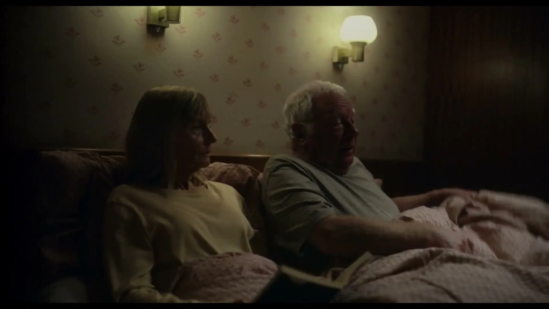 A middle-aged woman and an elderly man lie in bed, covered by blankets, as the man appears to be talking in a dimly lit bedroom.