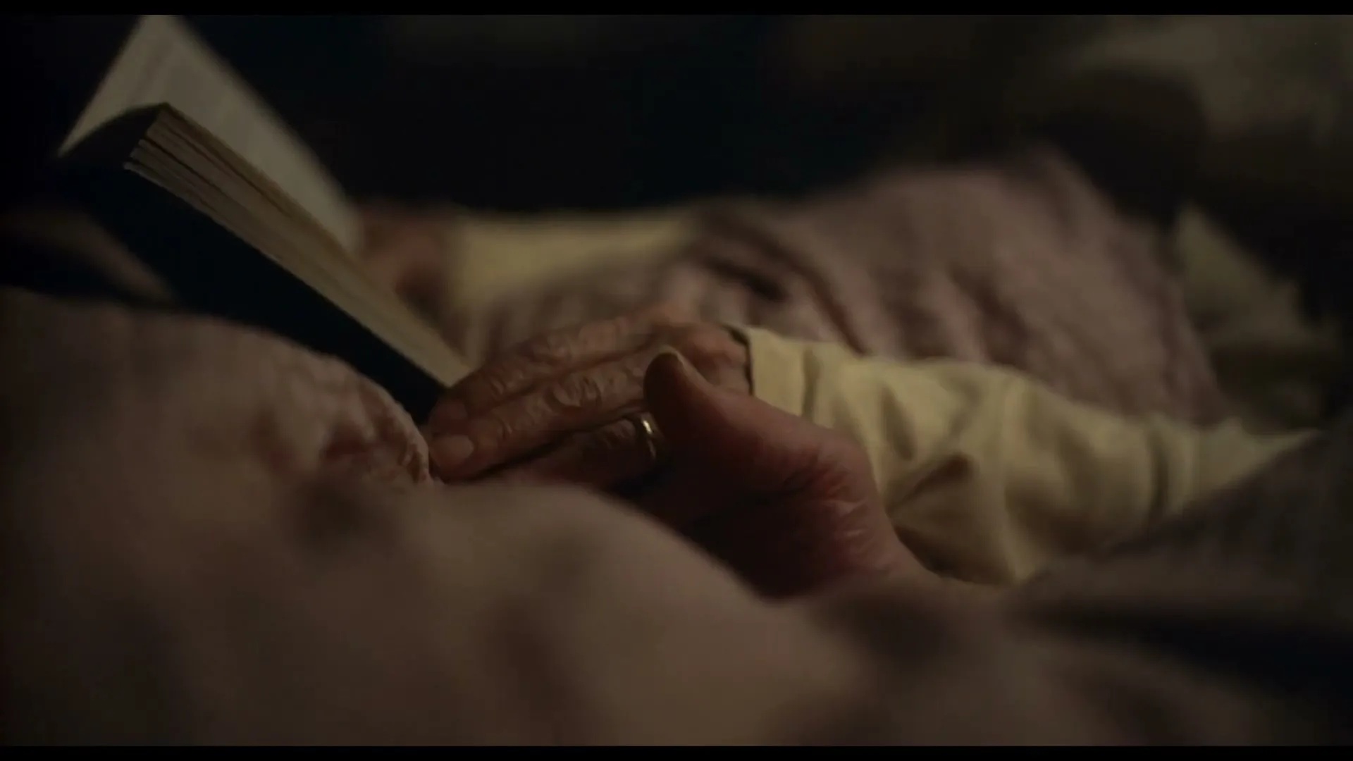 A close-up shot shows an elderly person's hand, wearing a wedding ring, resting on a book in bed, then another hand gently covers it.