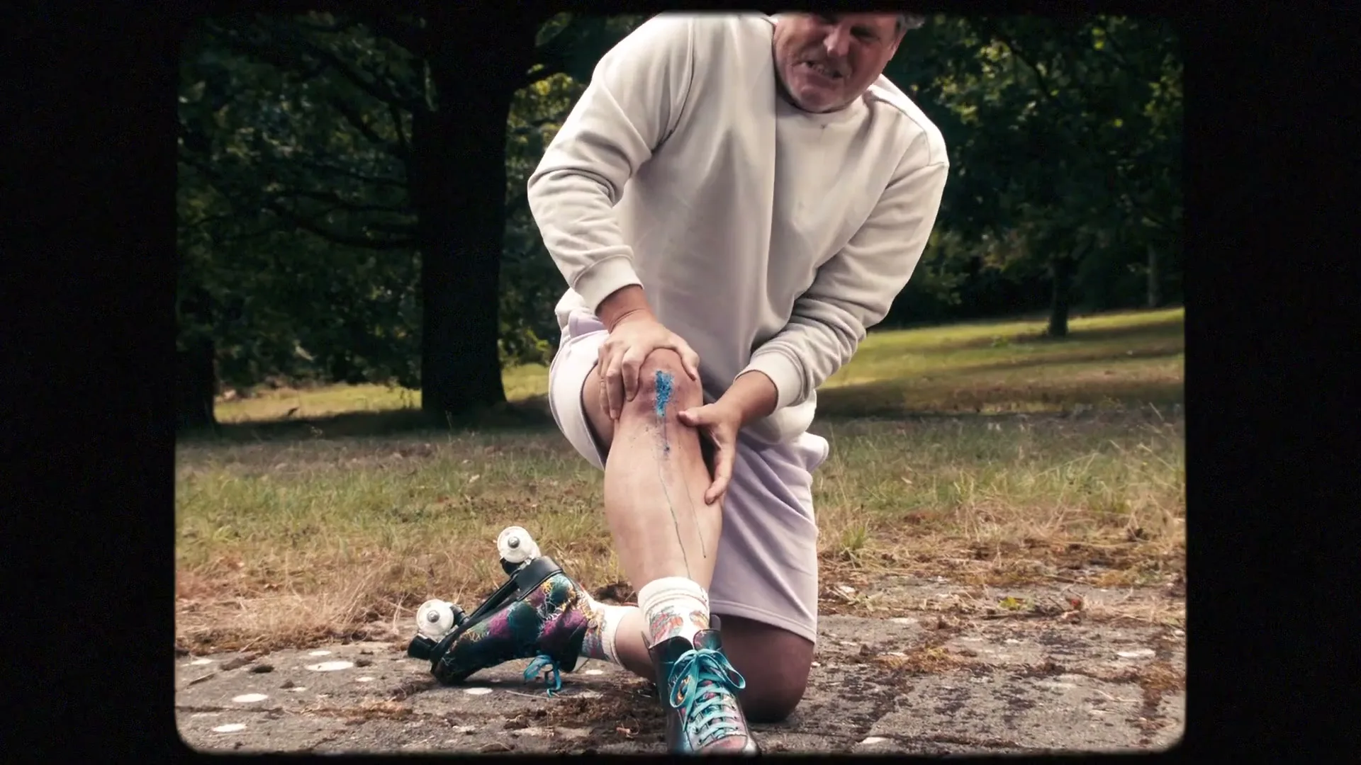 A middle-aged man on roller skates kneels on the ground, clutching his knee as blue liquid oozes from a wound in a park.