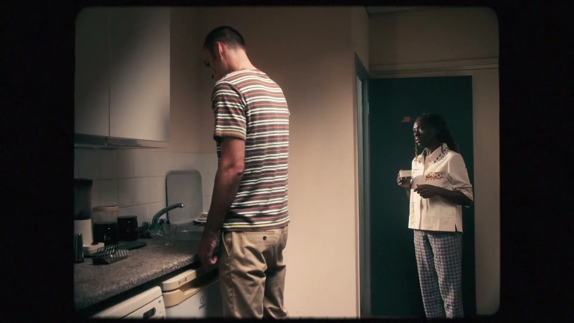 A man stands at a kitchen counter, back to the camera, as a woman enters the room holding a mug, looking at him with concern.